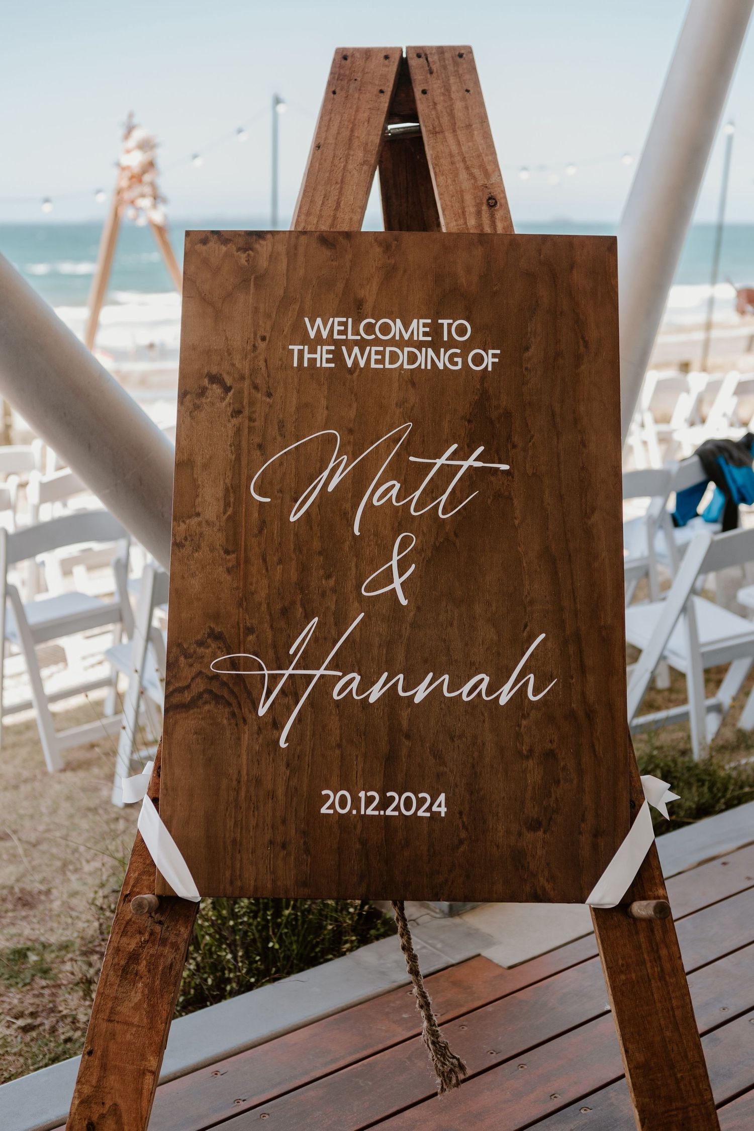 Wooden welcome sign for Matt and Hannah's beach wedding displayed on an easel with ocean waves in the background. Papamoa Surf Lifesaving Cl