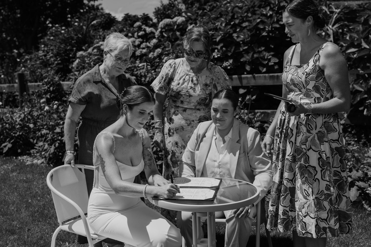 Black and white photo of a group of women gathered around a garden table, examining documents outdoors.
