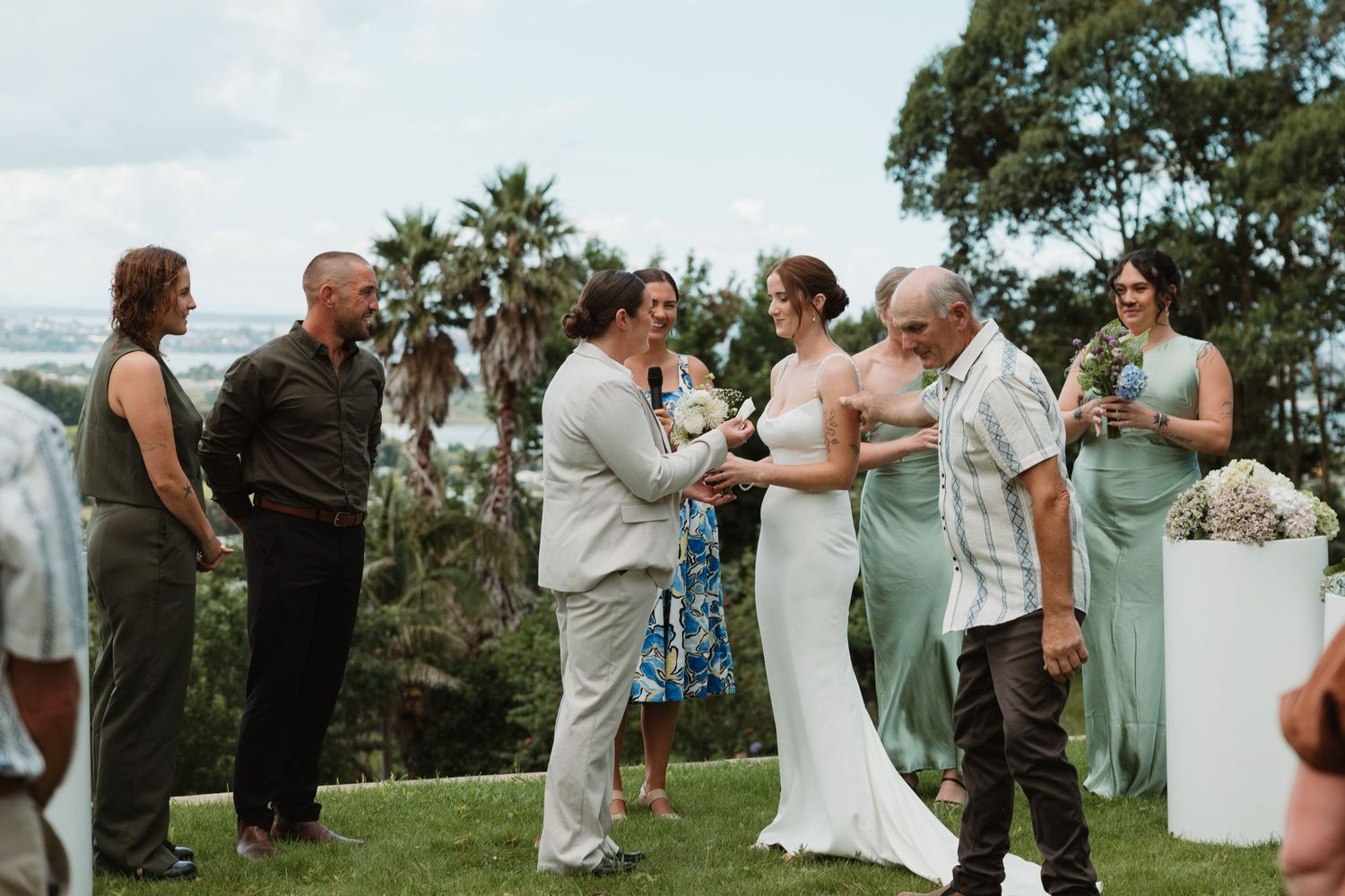 Bride and groom exchange vows outdoors at a garden wedding ceremony with guests and bridesmaid in sage green dress.