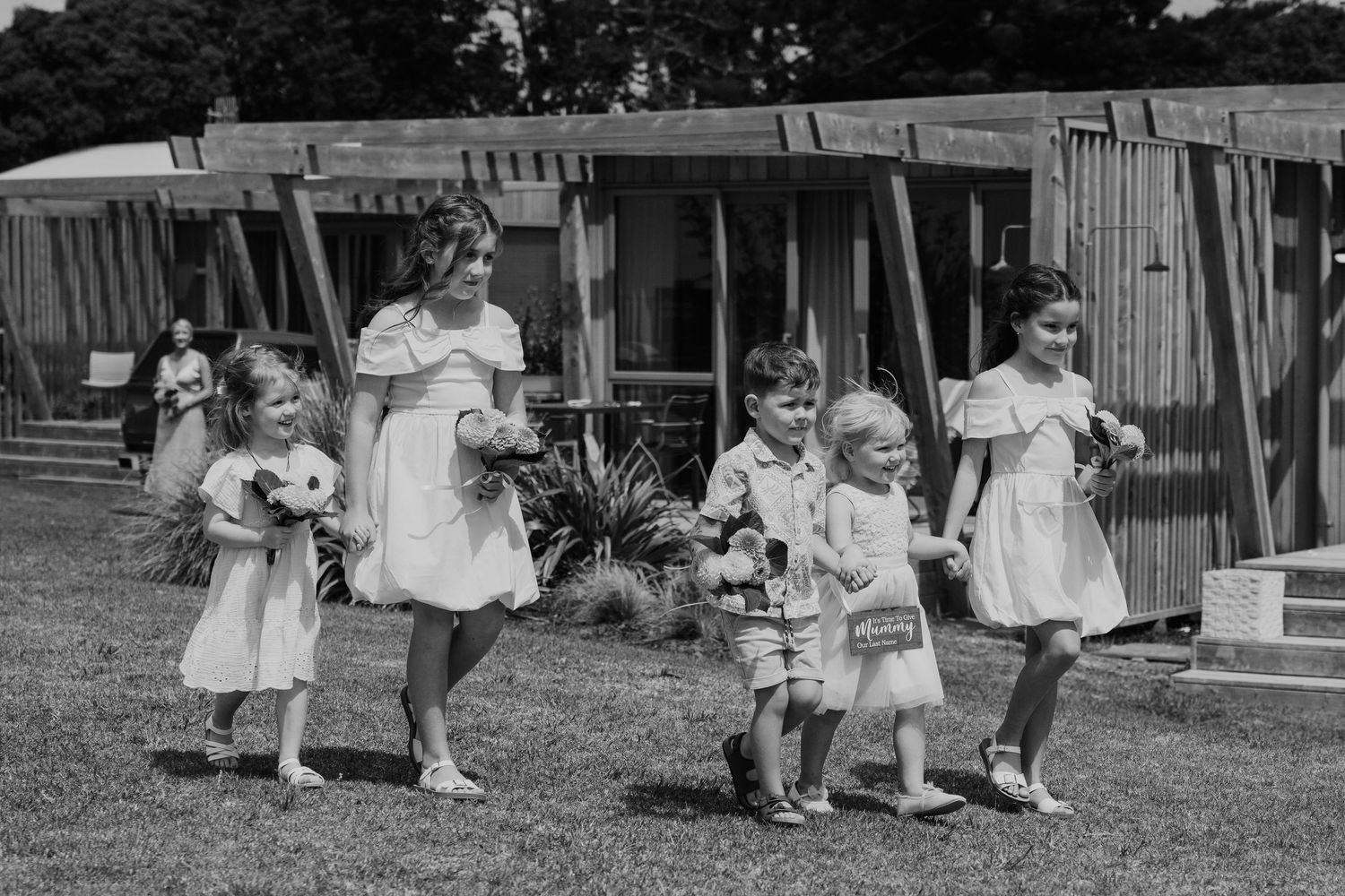 Black and white photo of children in white dresses walking on grass near wooden buildings at an outdoor event.