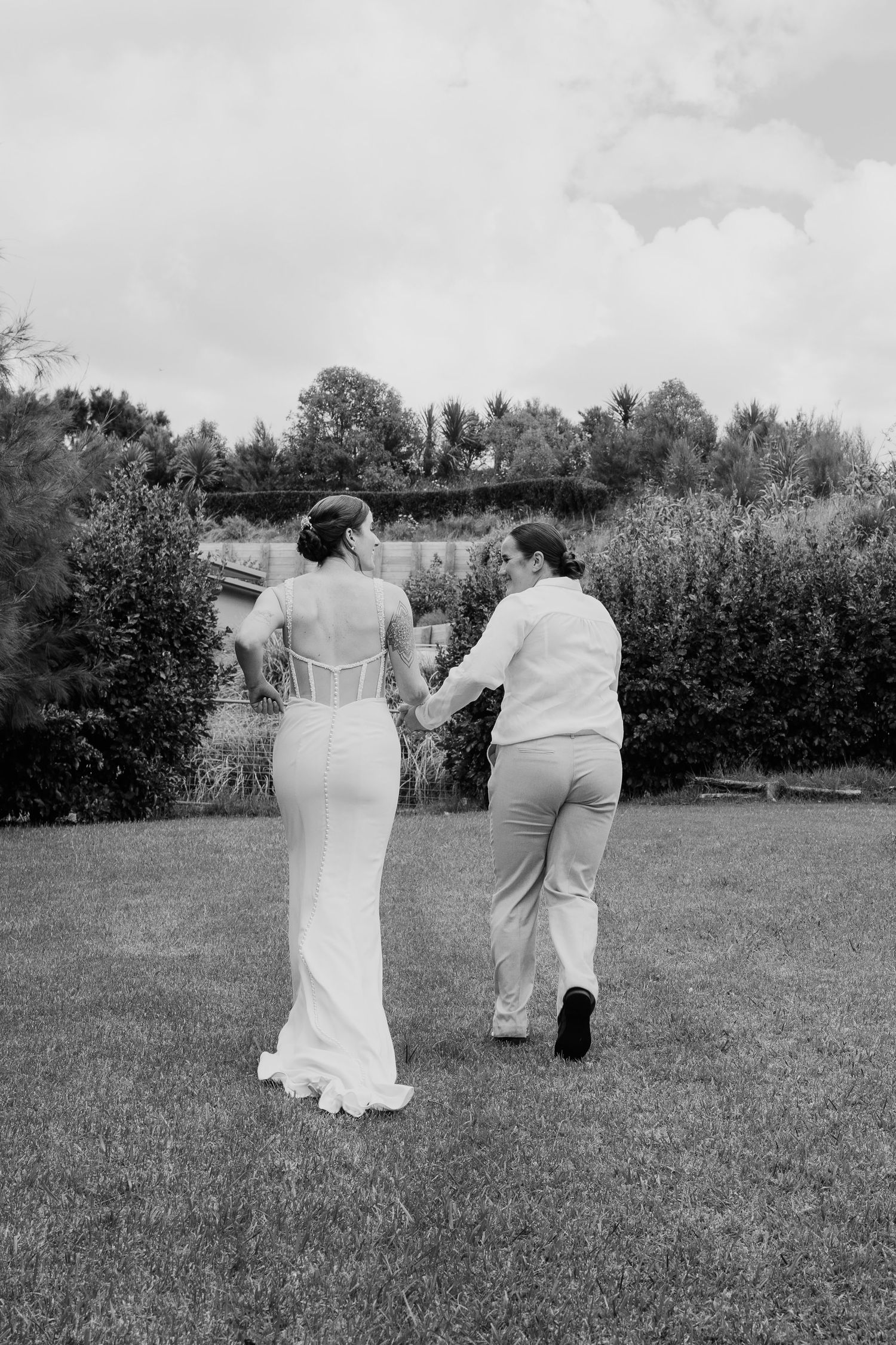 Black and white photo of a bride and partner walking hand in hand across a lush garden lawn on their wedding day.