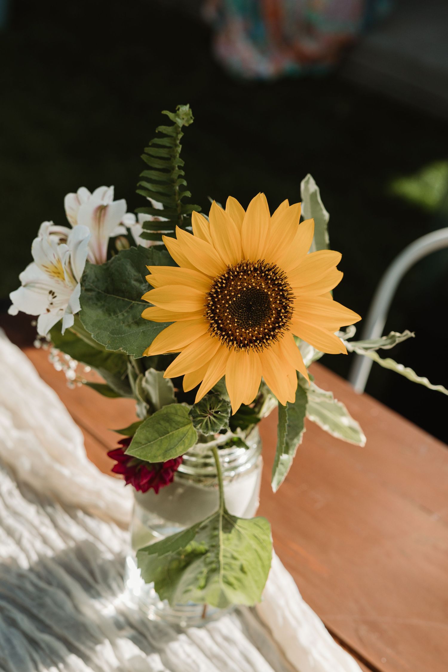 Bright sunflower arrangement in a mason jar with white alstroemeria and ferns on a wooden table.