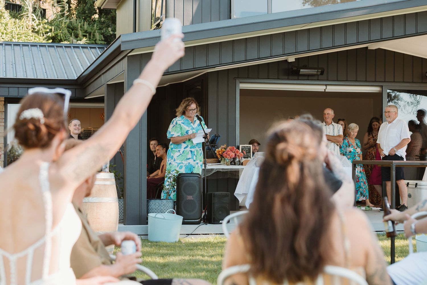 Wedding toast outdoors with guests raising glasses as a woman in floral dress speaks at a backyard reception.