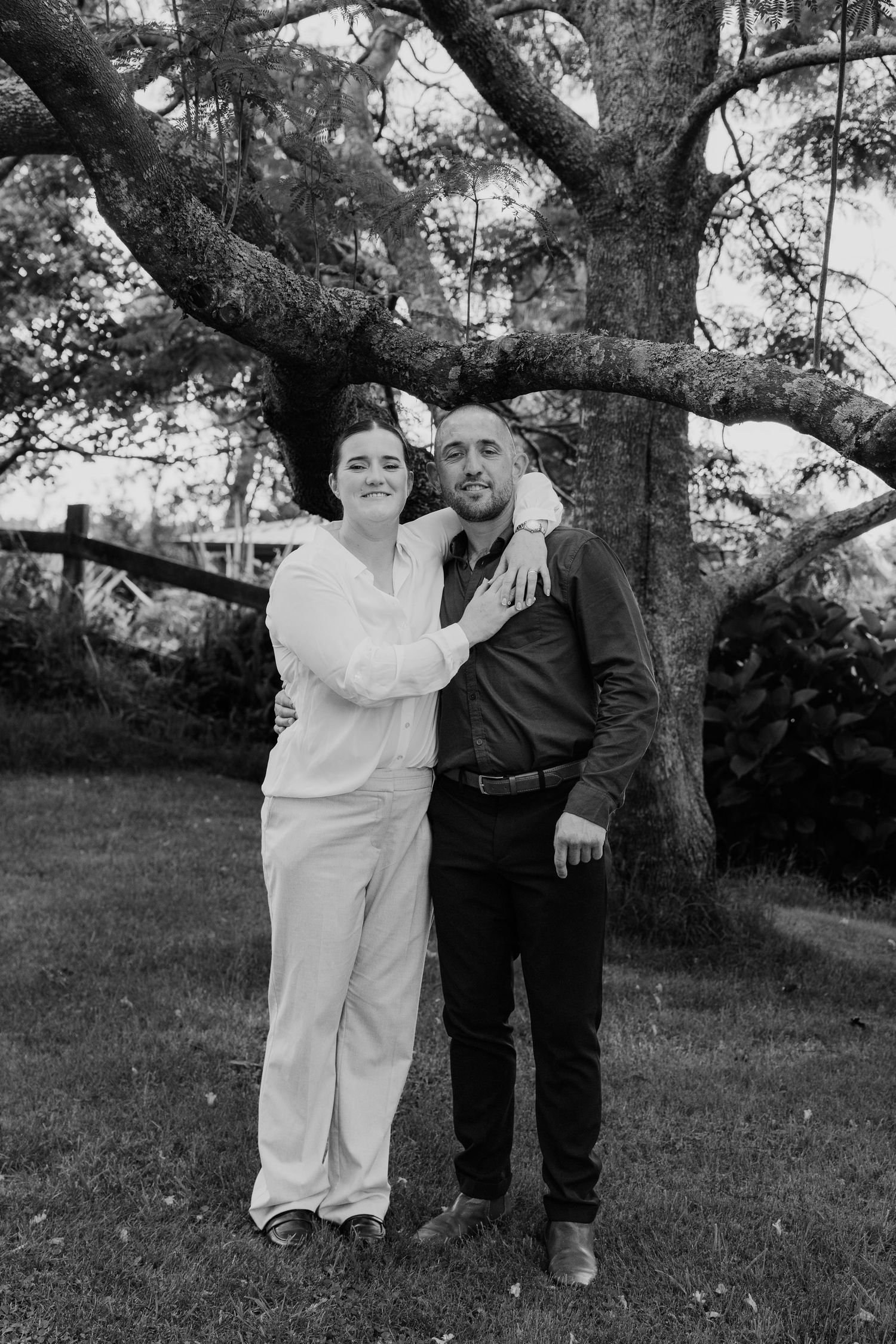 Black and white photo of a smiling couple embracing outdoors under a large tree on a green lawn.