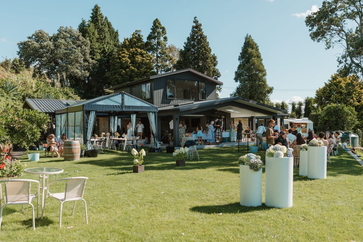 Outdoor wedding reception on lush green lawn with elegant white decor, floral arrangements, and a modern venue surrounded by trees.
