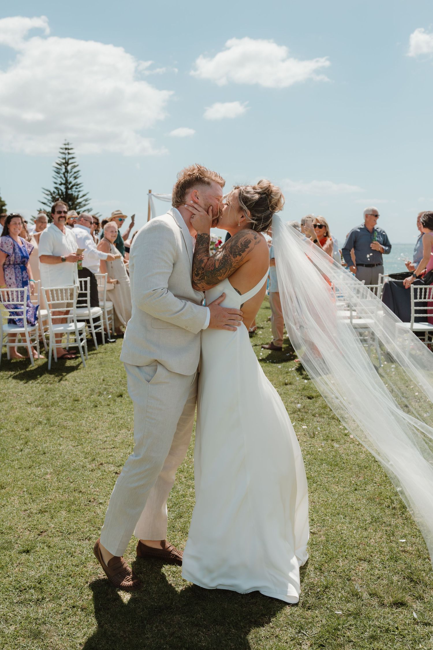 Bride and groom share first kiss at outdoor wedding ceremony on green lawn under blue sky with guests seated in white chairs.