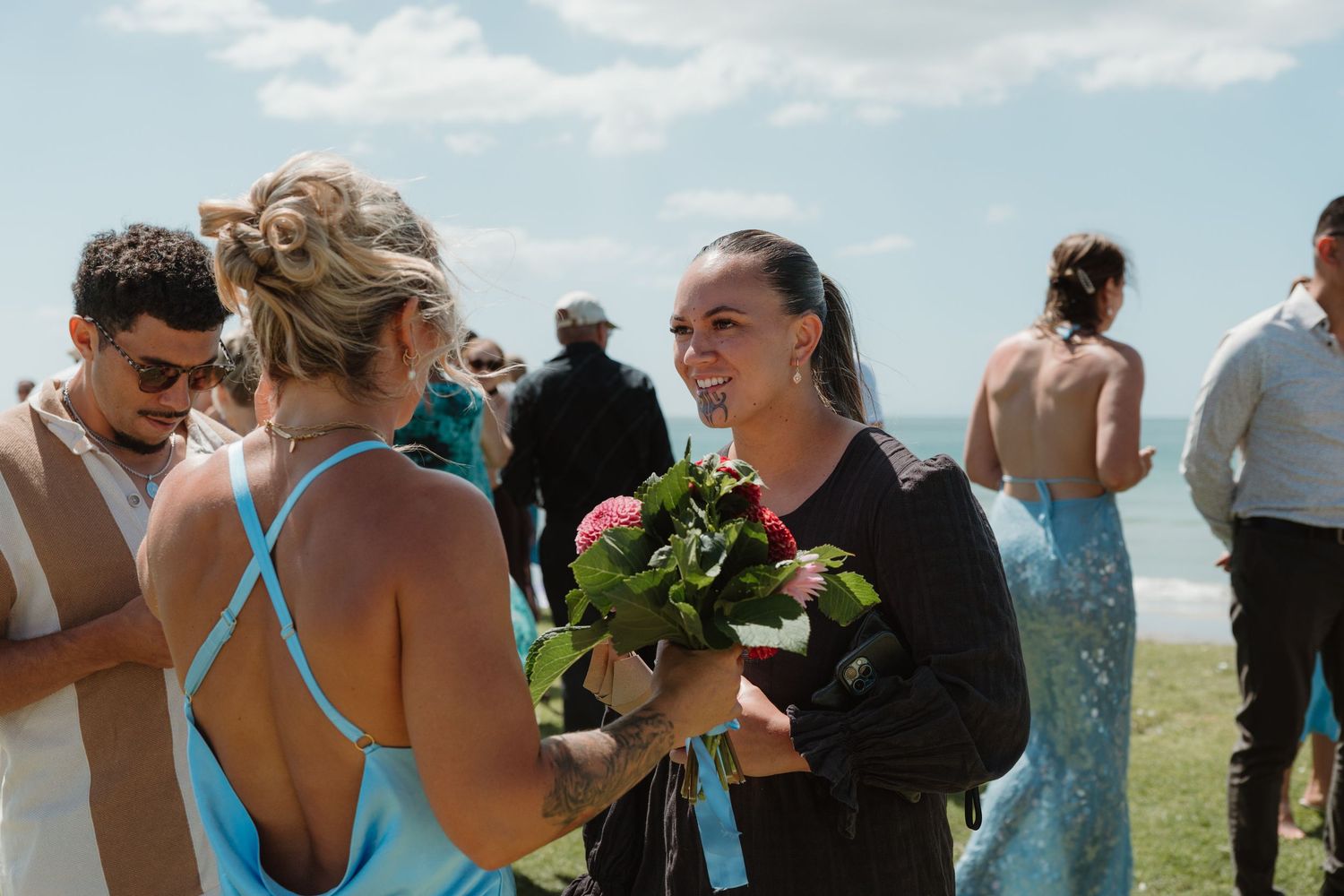 Women in blue dress holding bouquet chat at outdoor coastal wedding celebration on sunny day.