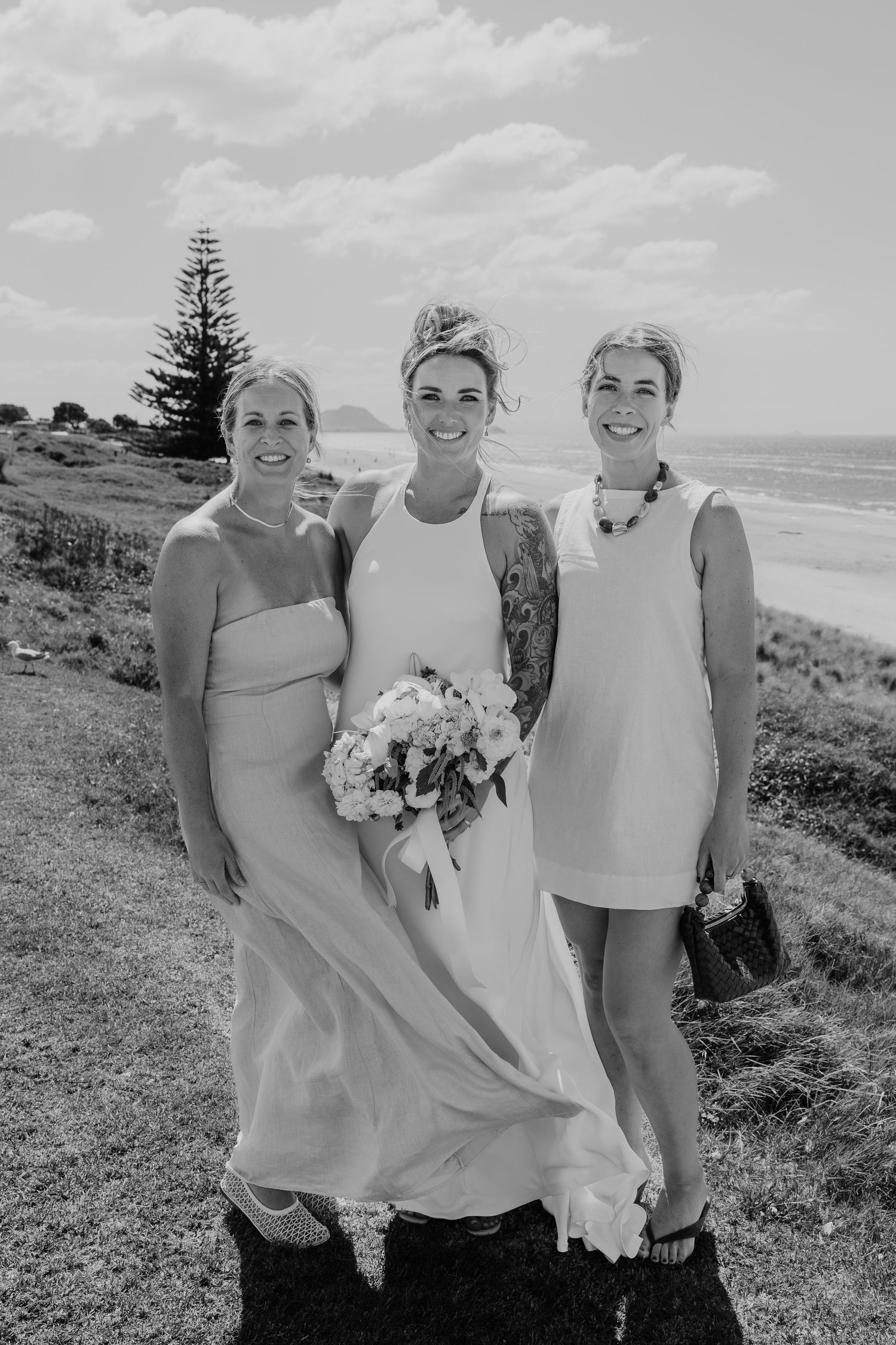 Black and white photo of a bride holding bouquet flanked by two women, posing outdoors near a scenic beach.