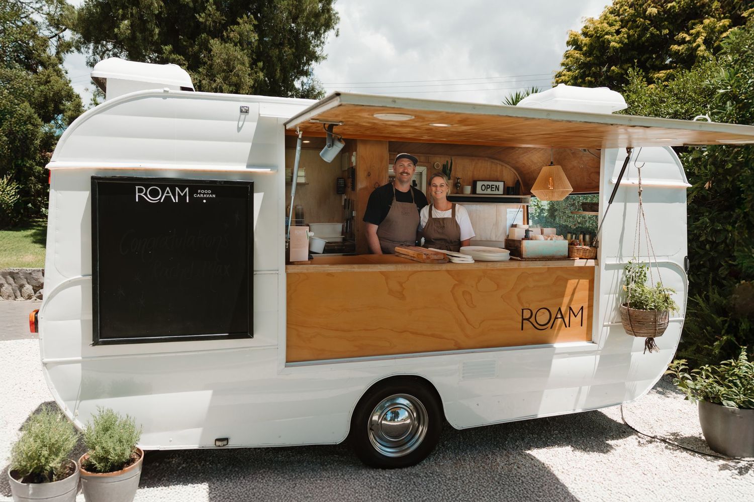Two vendors smile inside a white ROAM food truck trailer with wooden accents, serving customers outdoors.