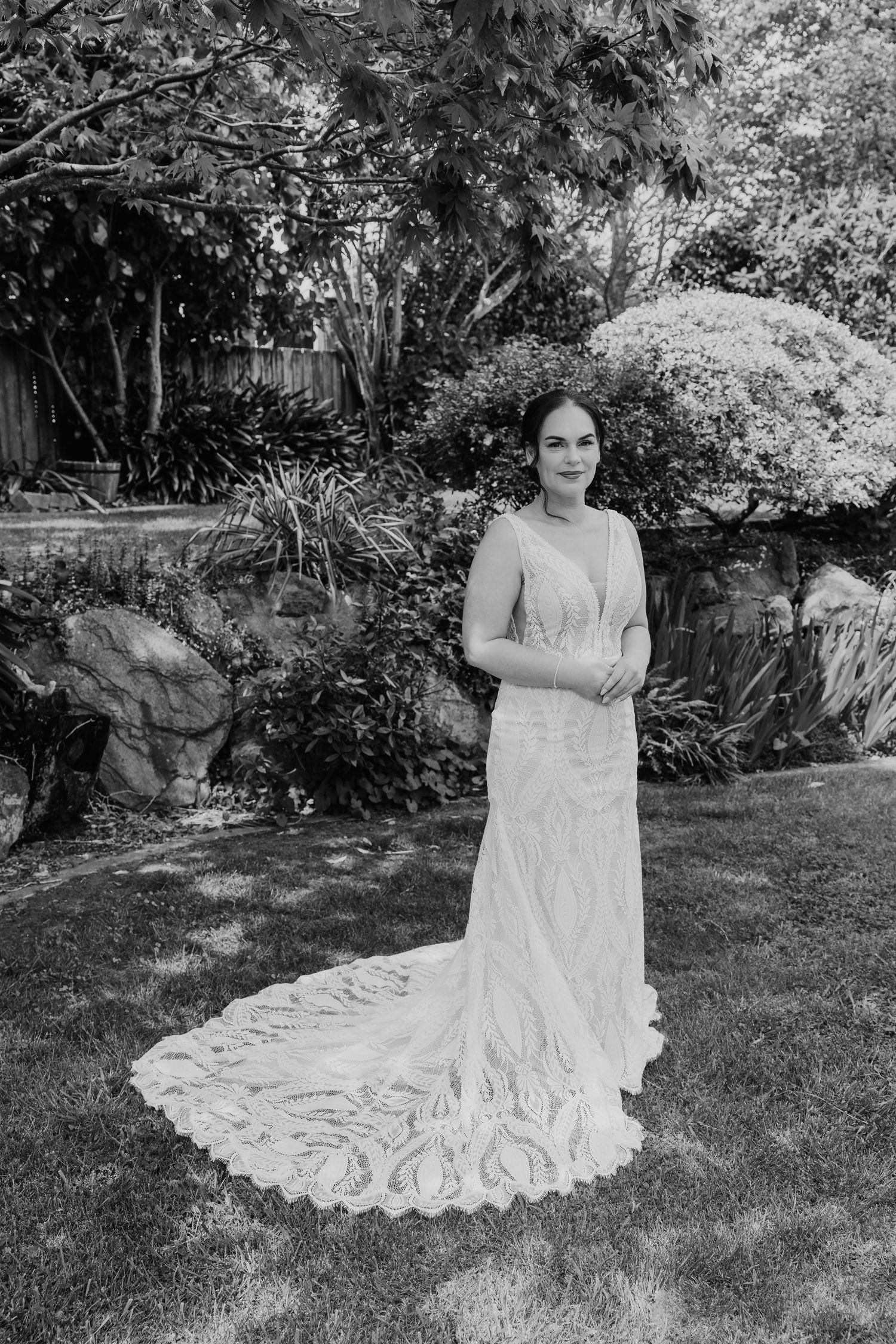 A person wearing a flowing white lace wedding dress poses in a garden setting with natural lighting and greenery.