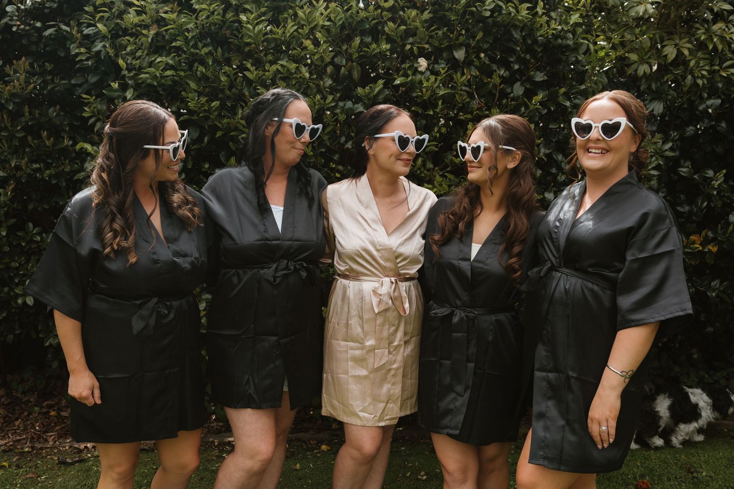 A group of five friends wearing black robes and one in gold stand together outdoors smiling in sunglasses.