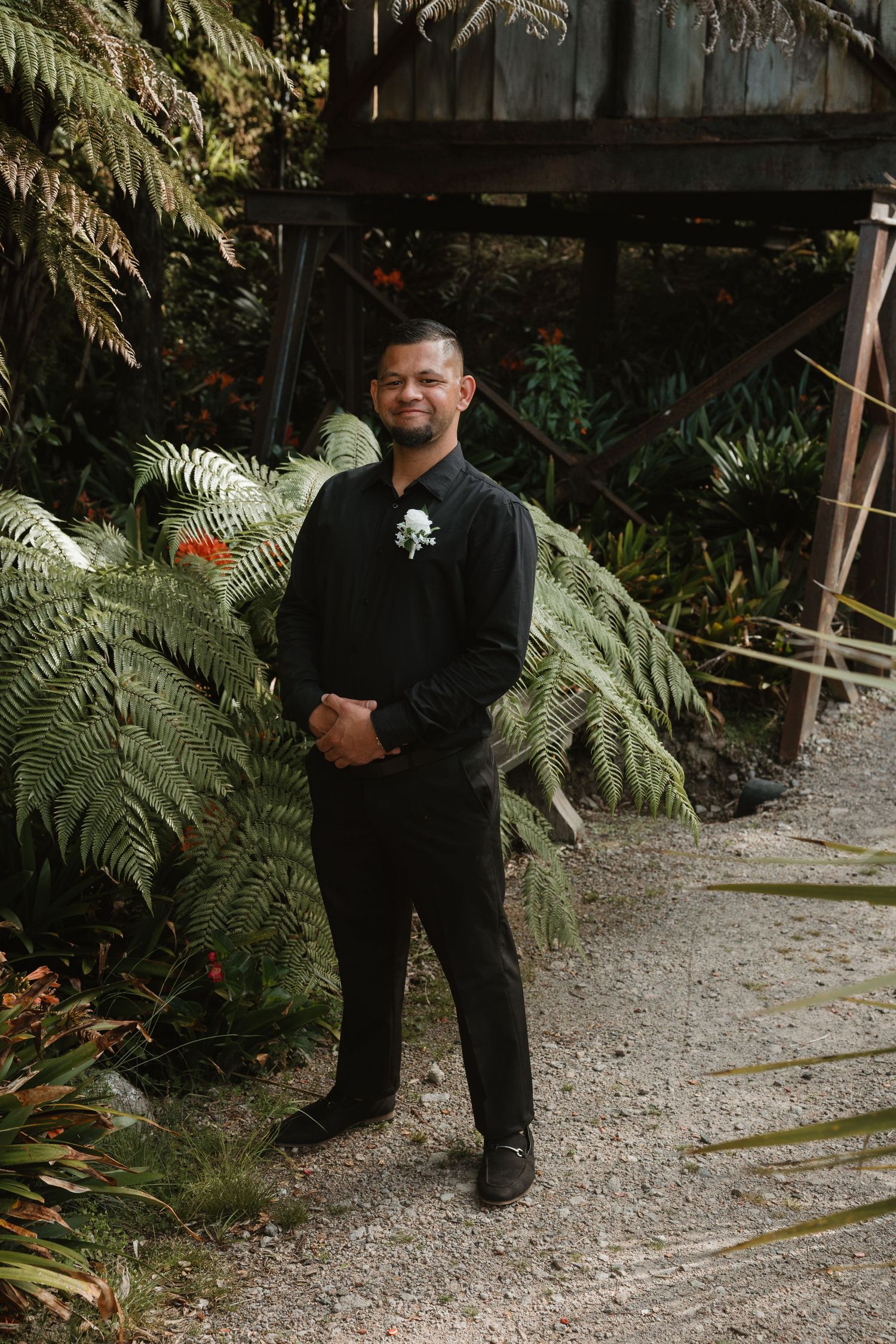 Person wearing black clothing stands on gravel path surrounded by tropical ferns and palm fronds in garden setting.