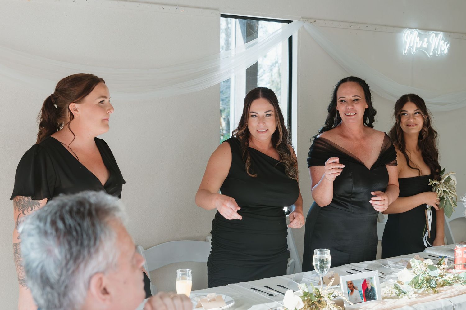 Wedding party members in elegant black dresses share a joyful moment during reception speeches at a decorated table.