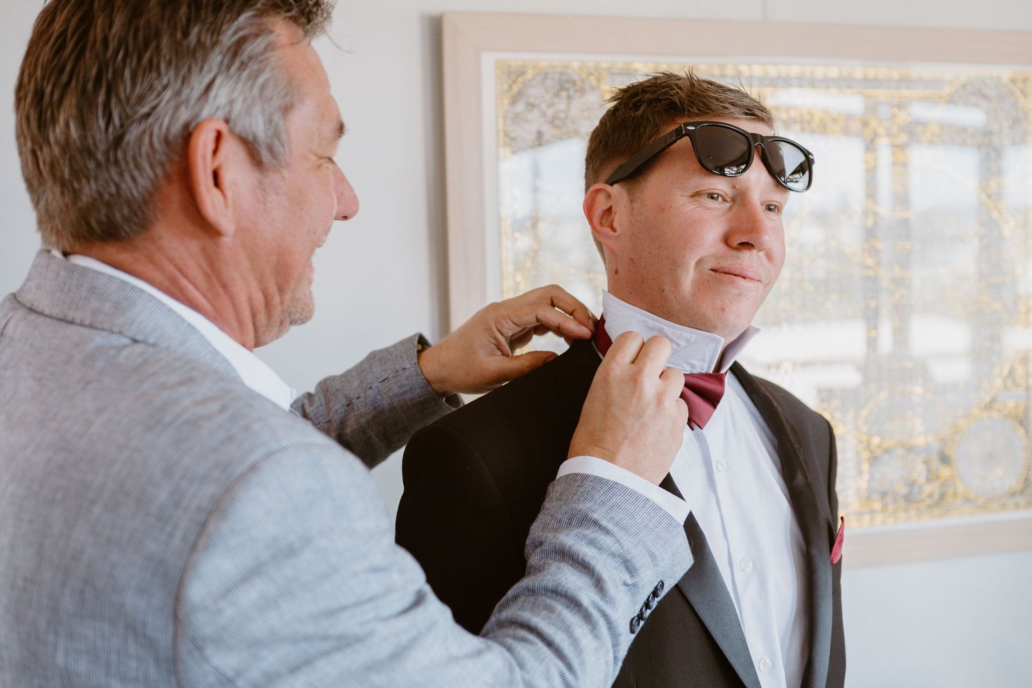 Two people sharing a moment as one adjusts the other's formal attire in front of a window.