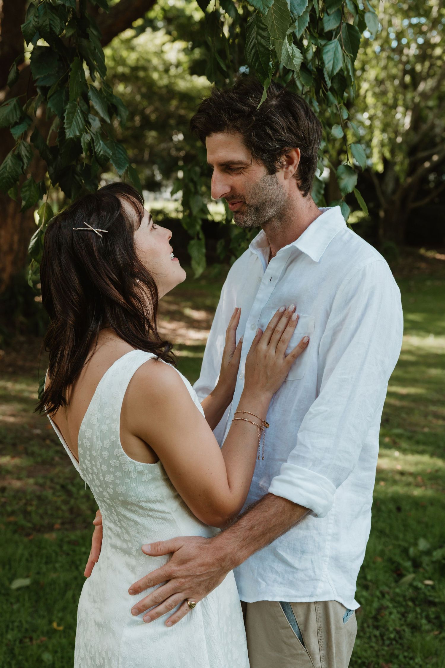 A couple in white attire shares a romantic embrace outdoors, surrounded by lush green trees in a garden setting.