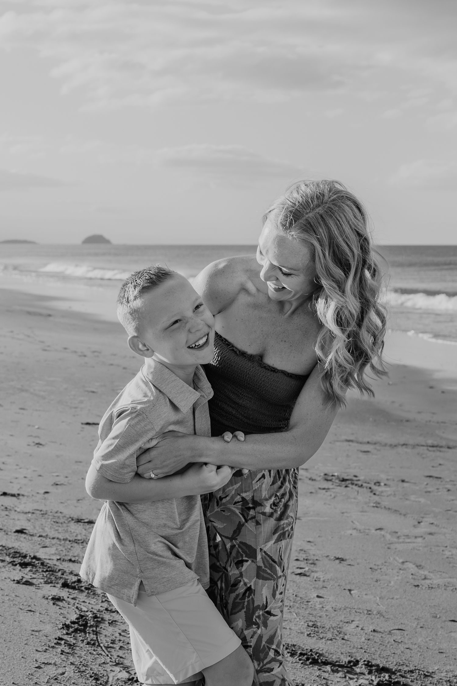 Black and white photo of a laughing mother hugging her young son on a sandy beach with ocean waves behind them.