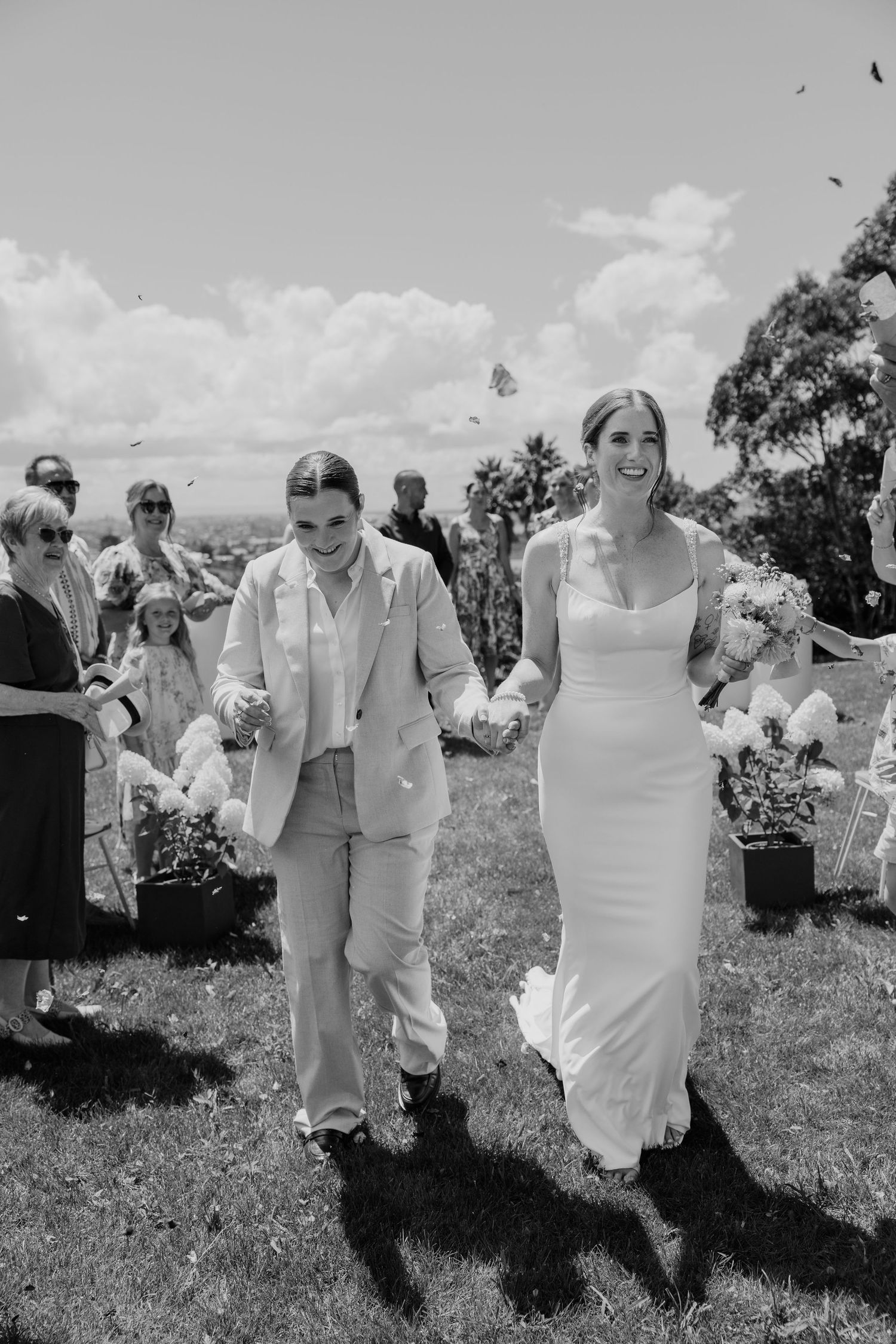 Two brides walk hand-in-hand down the aisle after their outdoor wedding ceremony in black and white photo.