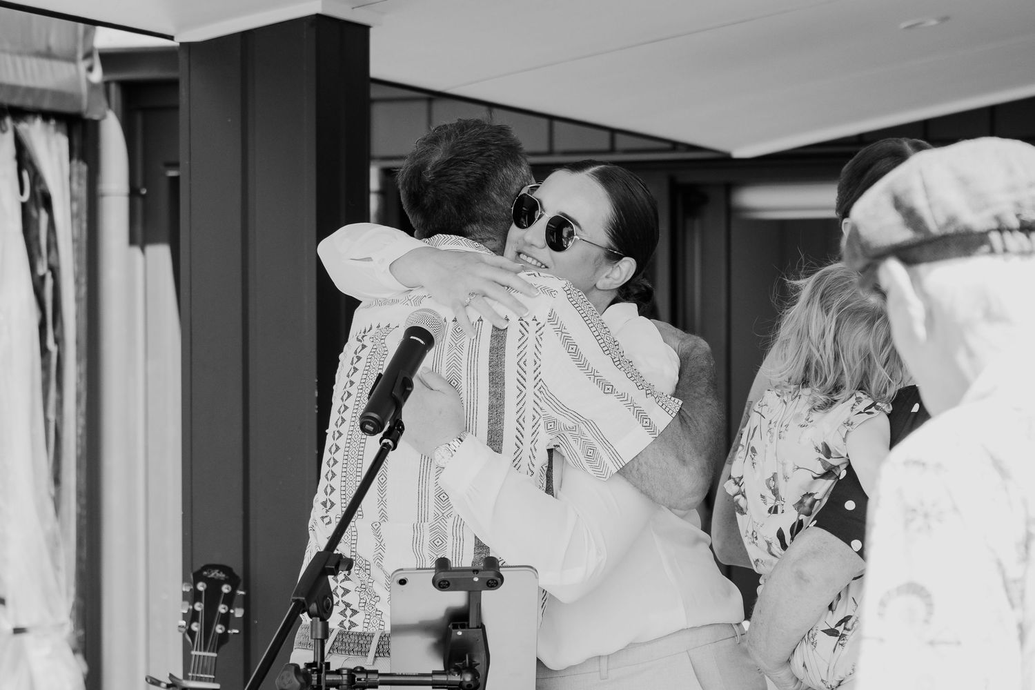Two people sharing a warm hug near a microphone stand and guitar in a black and white photo.