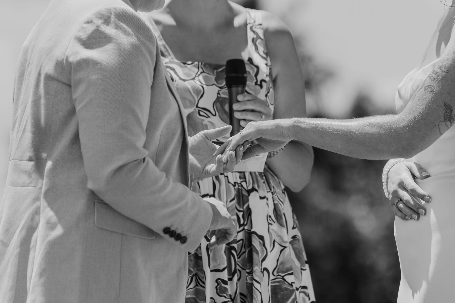 Black and white wedding ceremony photo of guests exchanging rings during an outdoor celebration.