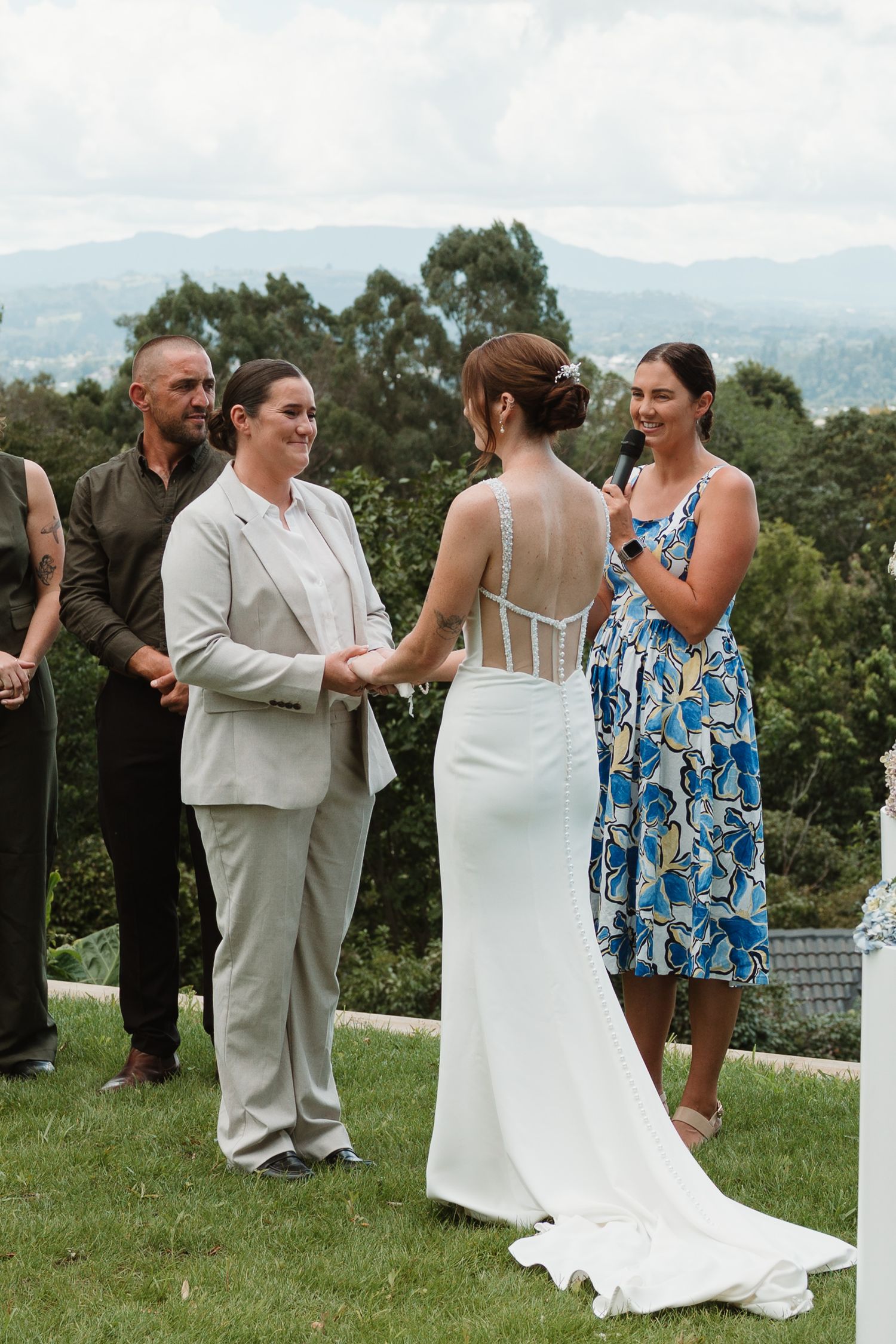 Two brides hold hands during an outdoor wedding ceremony, one in a white gown, the other in a beige suit.