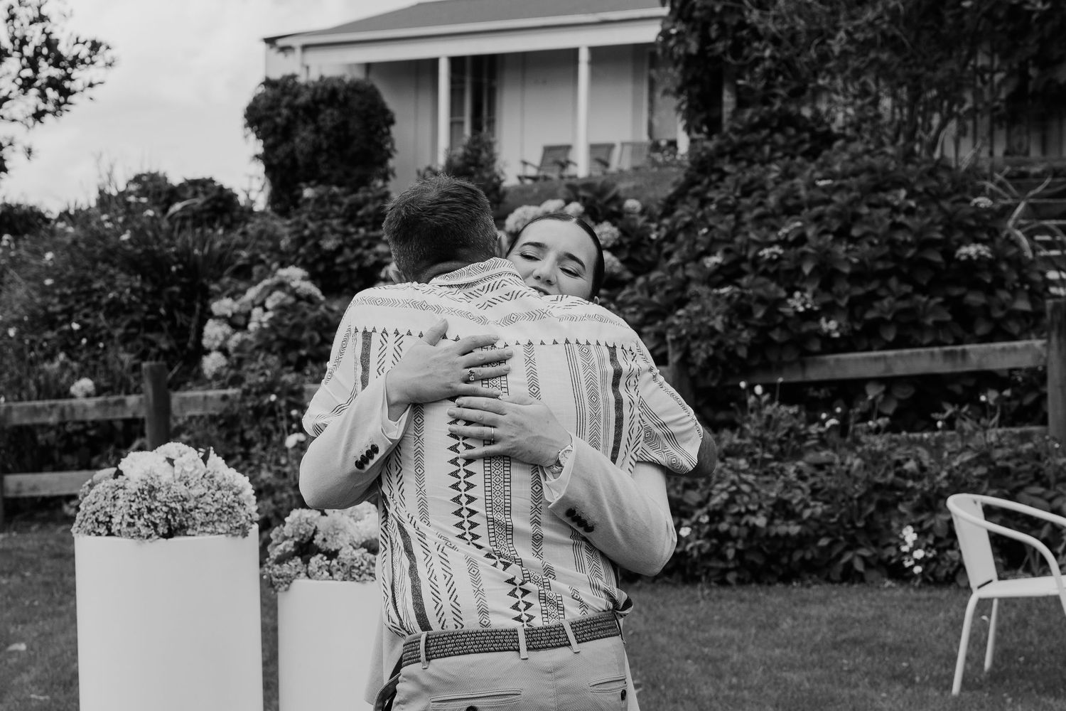 Black and white photo of a couple embracing in a garden with flowers and a house in the background.