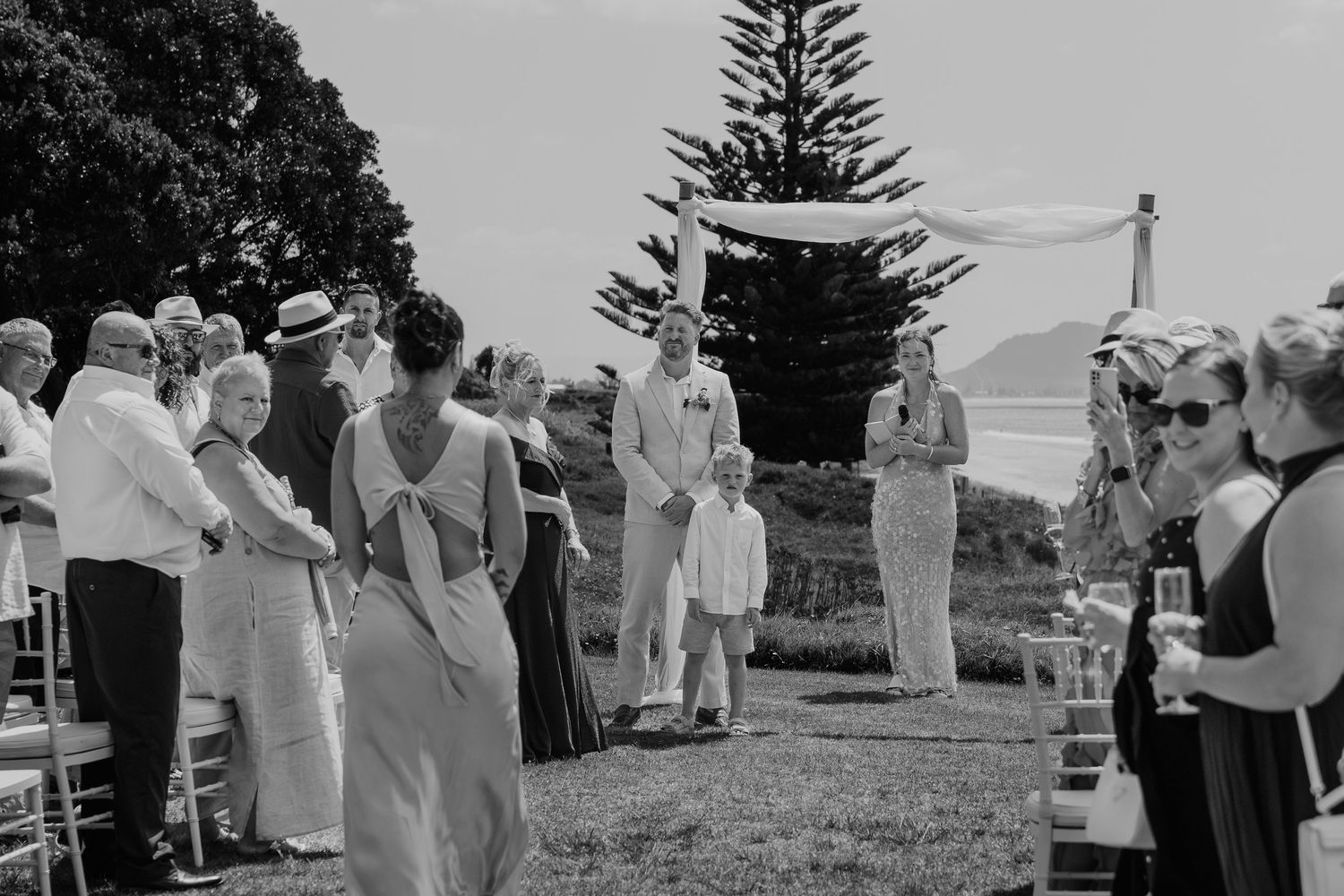 Black and white outdoor wedding ceremony on a lawn with guests seated, officiant speaking, and scenic ocean views.