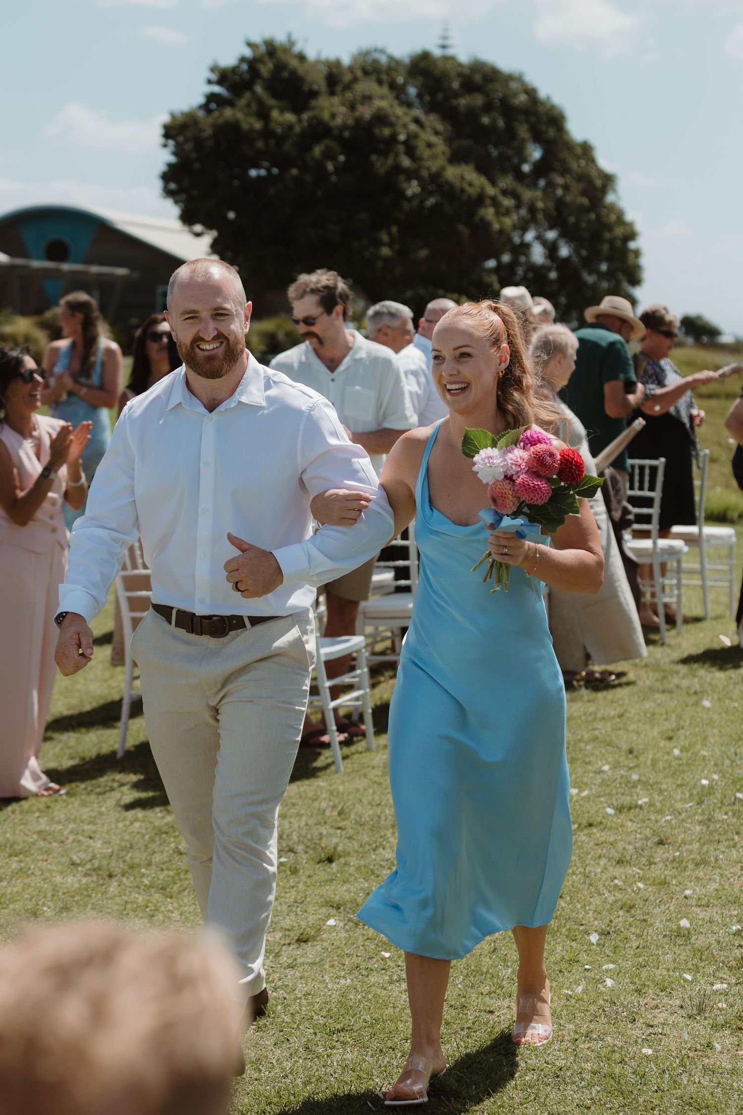 A joyful couple walks down the aisle at an outdoor wedding, woman in blue dress holding pink bouquet.