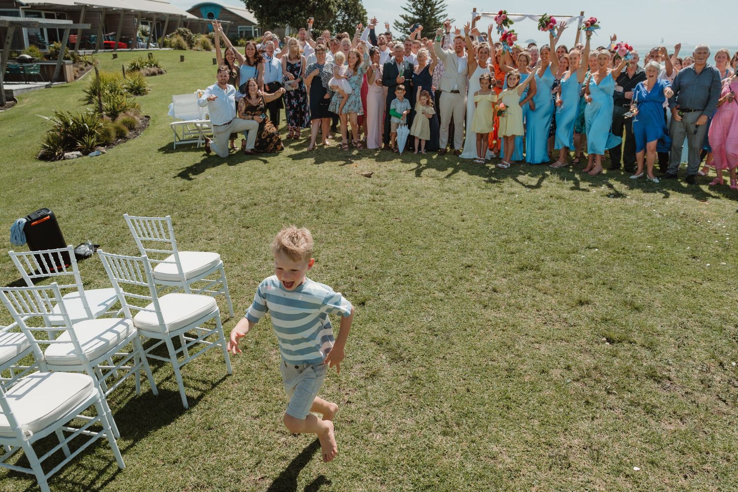 A young child runs across a lawn at an outdoor wedding while guests cheer and wave in the background.