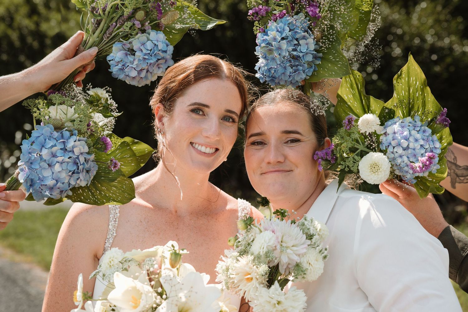 Two brides smile together surrounded by blue hydrangea and wildflower arrangements at their outdoor wedding ceremony.