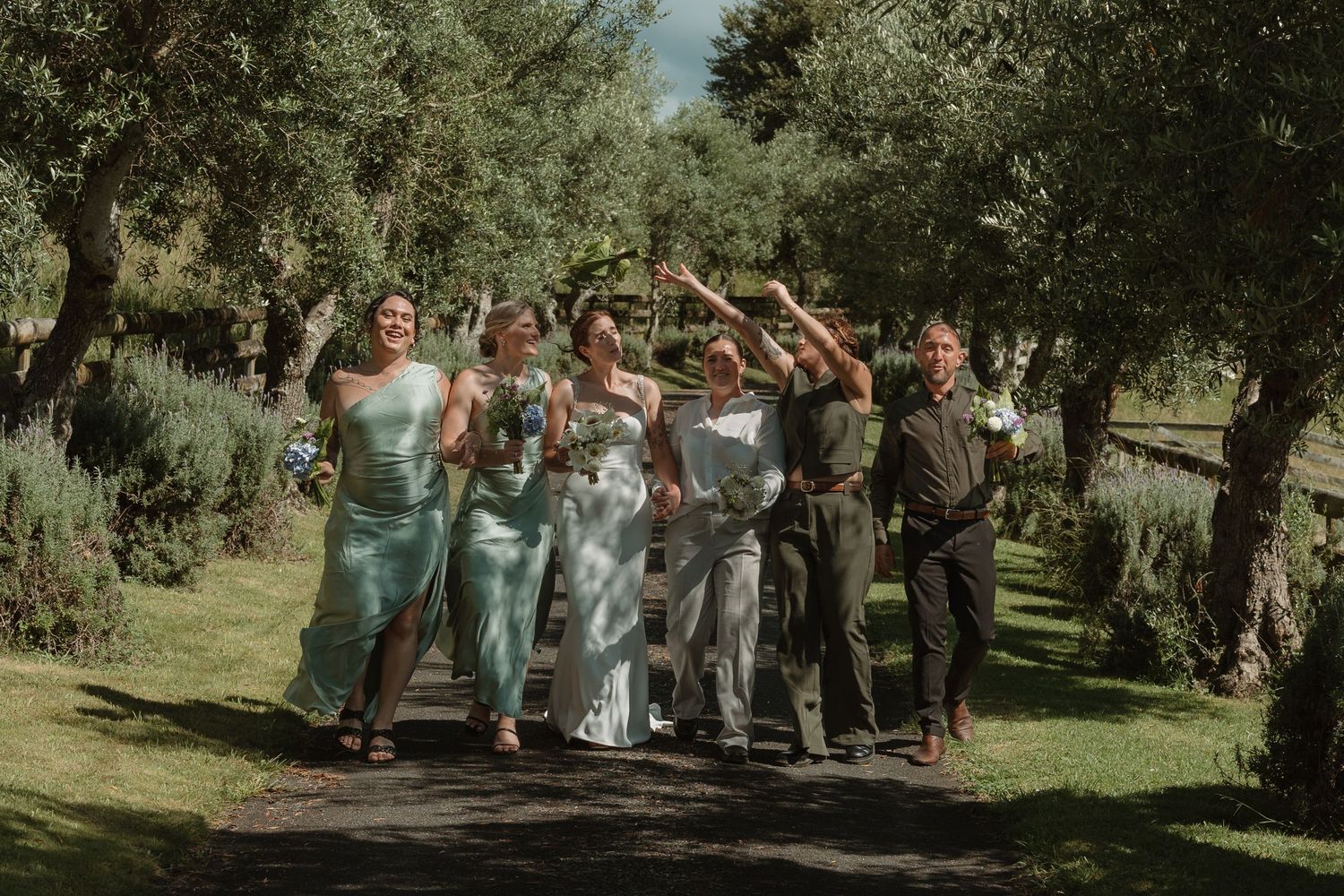 Bridal party in sage green dresses celebrating outdoors among olive trees at a sunny garden wedding.