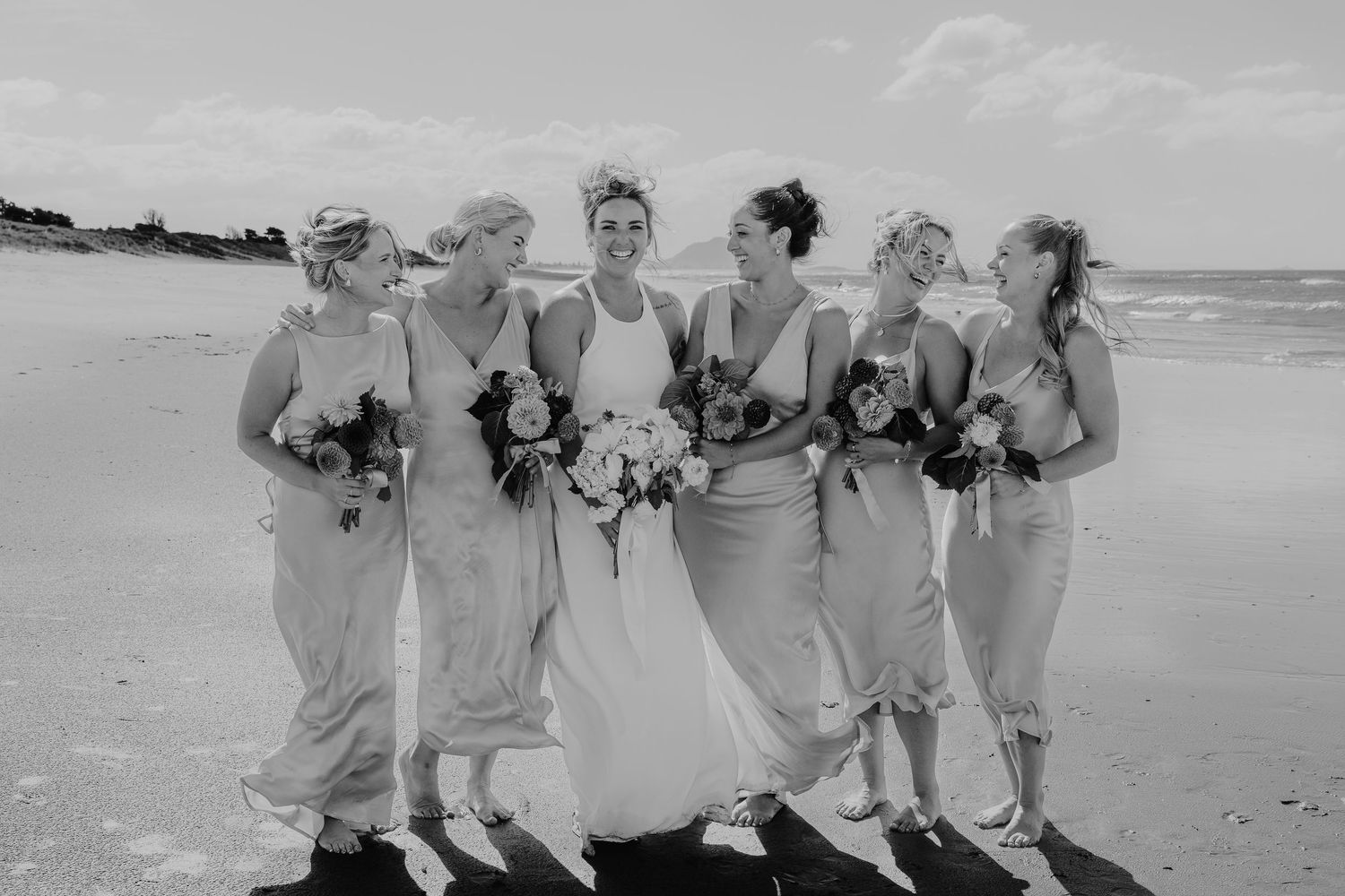 Bride and bridesmaids laughing together on a beach, holding bouquets, in elegant gowns, black and white wedding photo.
