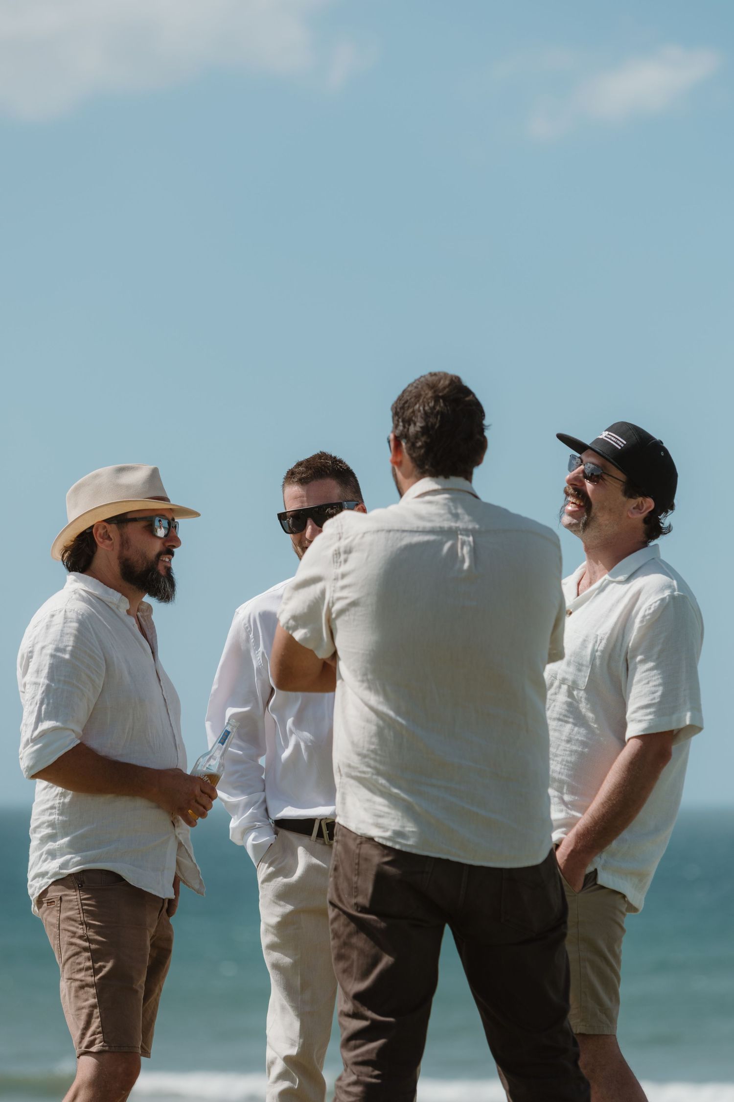 Four men in casual white shirts and hats laugh together on a sunny beach with ocean waves behind them.