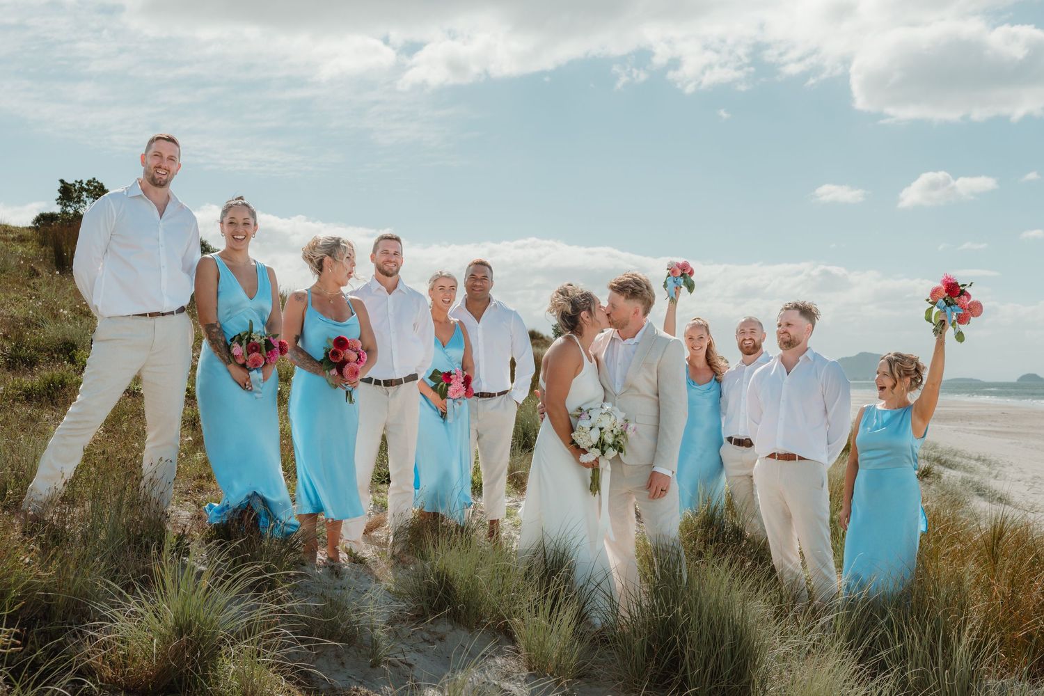 Beach wedding party in turquoise dresses and white shirts, bride and groom kissing on sandy dunes with ocean backdrop.