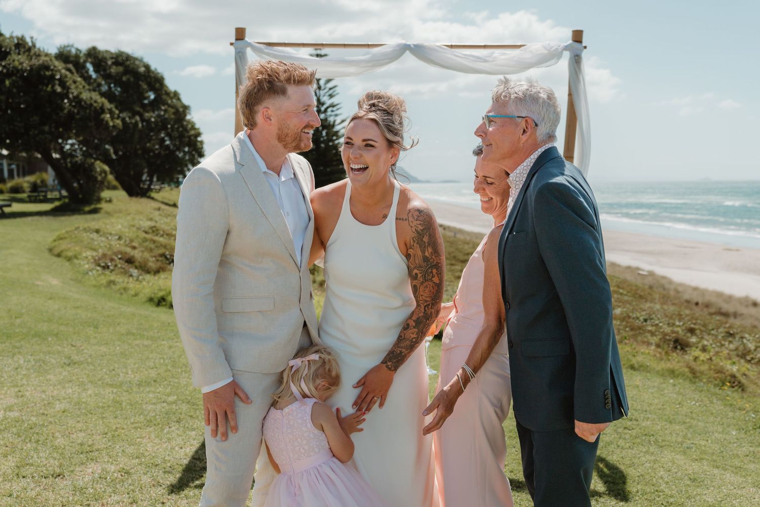 Outdoor beach wedding ceremony with bride, groom, and guests laughing joyfully on green lawn with ocean backdrop.