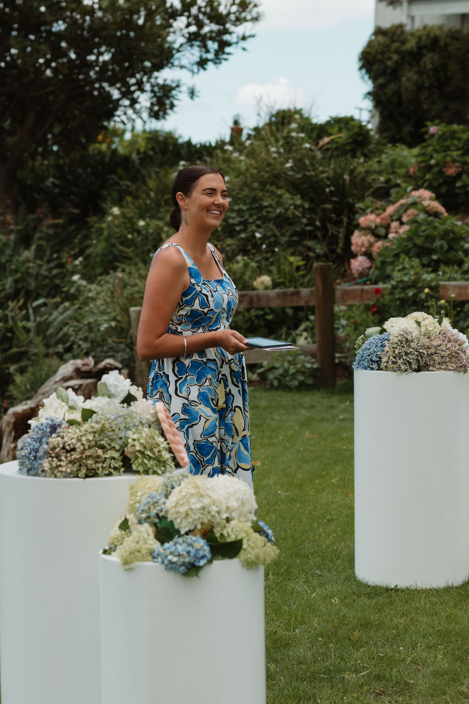 Woman in blue floral dress smiling at outdoor garden ceremony with white pedestals and hydrangea floral arrangements.