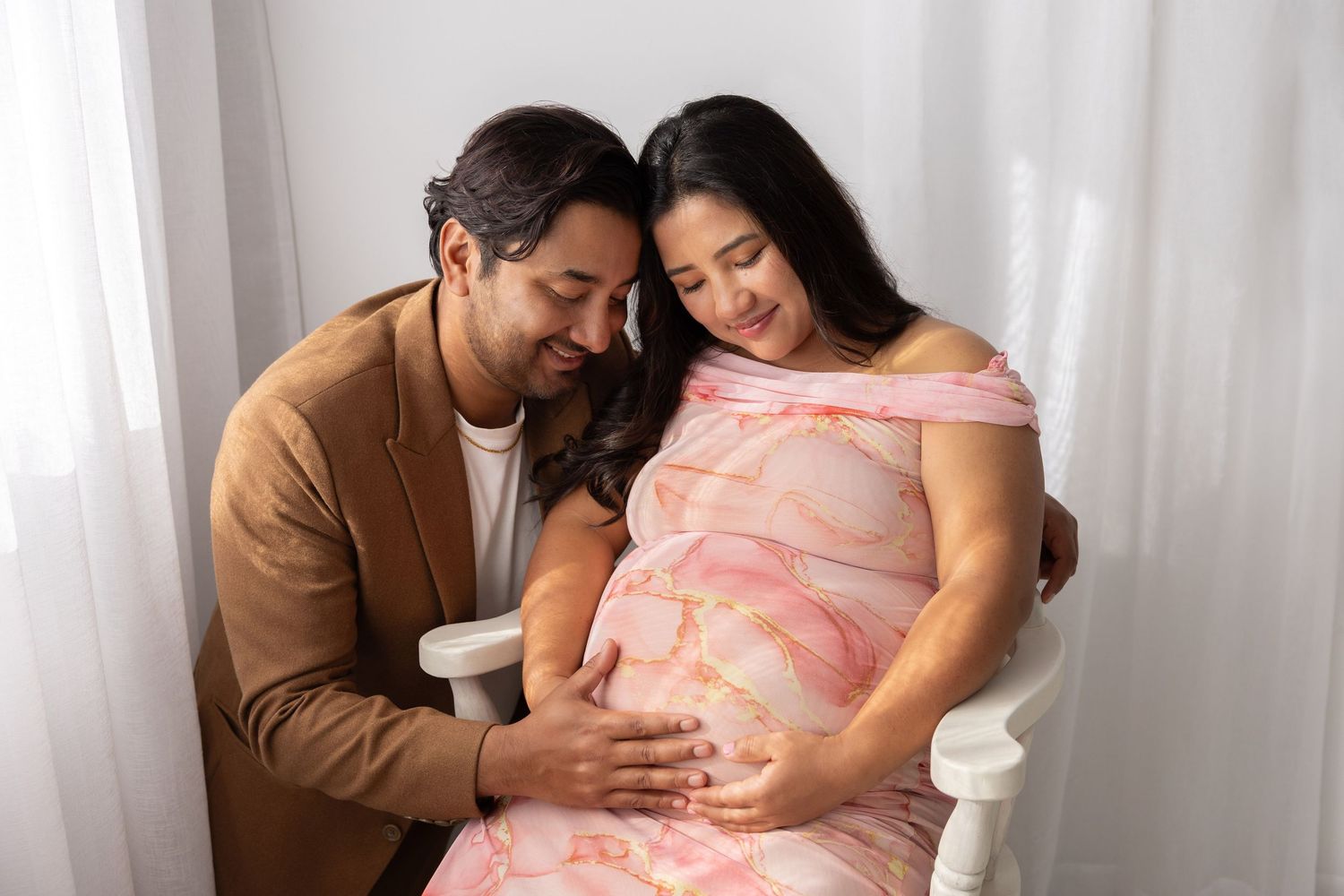 Couple embraces lovingly during an intimate maternity photo session, dressed in soft peach and brown tones.