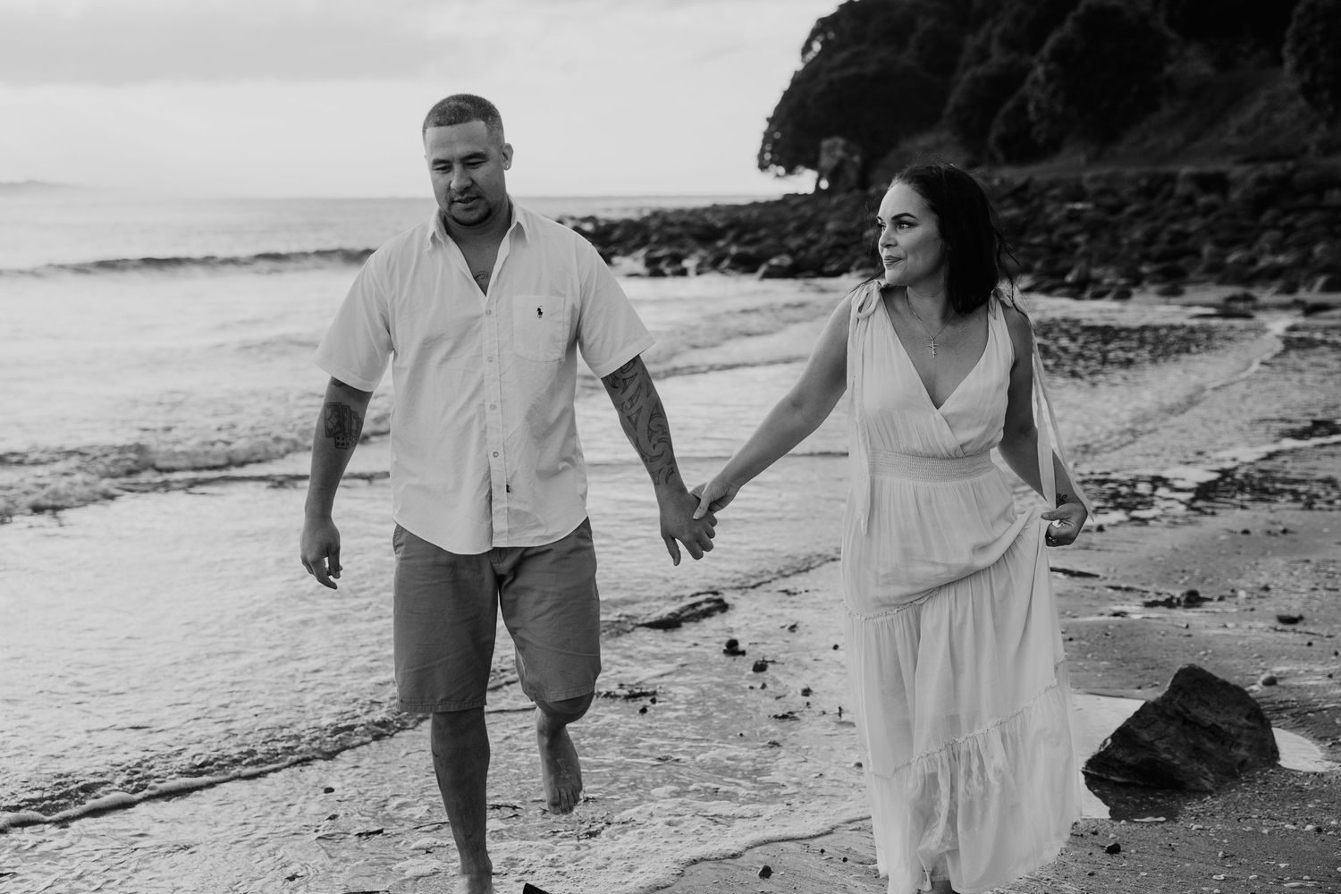 A couple in white outfits walks hand in hand along a rocky beach at sunset, creating romantic silhouettes against the ocean.