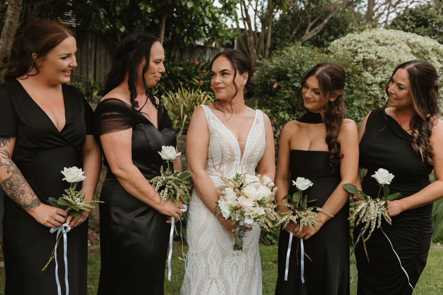 A bridal party in black dresses and white bouquets laughs together during outdoor wedding photos.