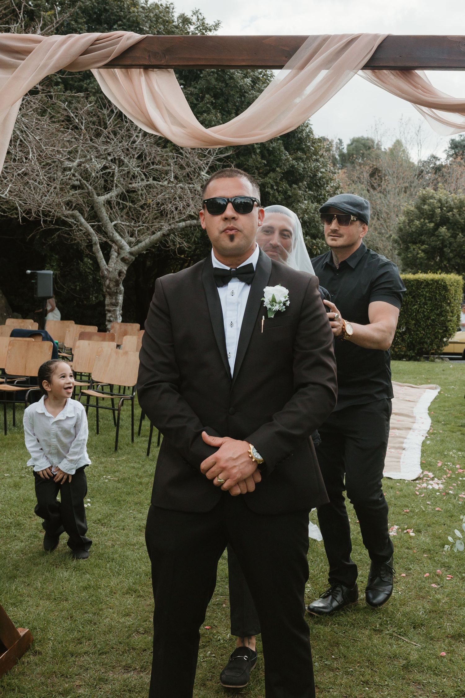 A group of formally dressed people in black suits and sunglasses stand together on a grassy lawn at an outdoor event.