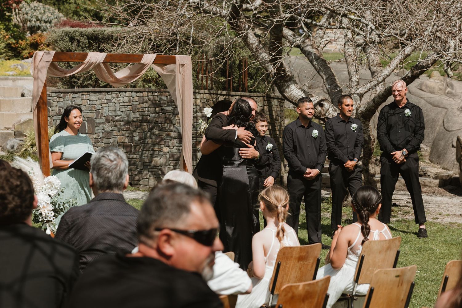 An outdoor wedding ceremony setup with wooden chairs and a draped arch against a stone wall in a garden setting.