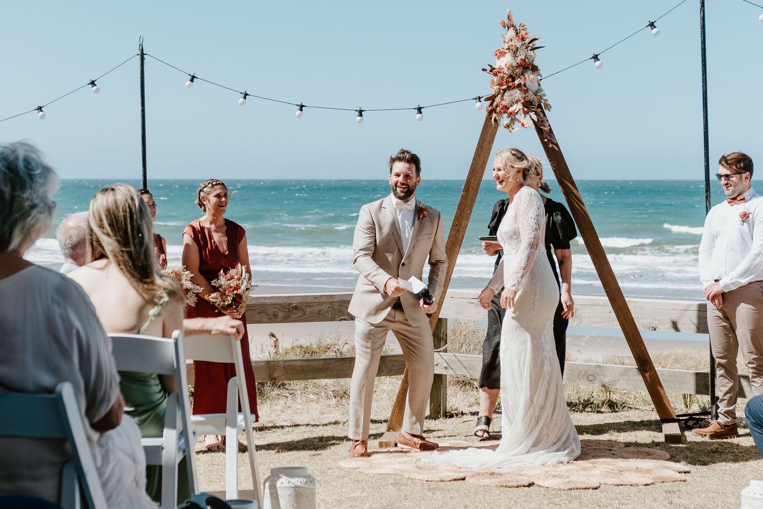 Beach wedding ceremony setup at sunset with wooden arch, string lights and chairs arranged on the sand overlooking the ocean.