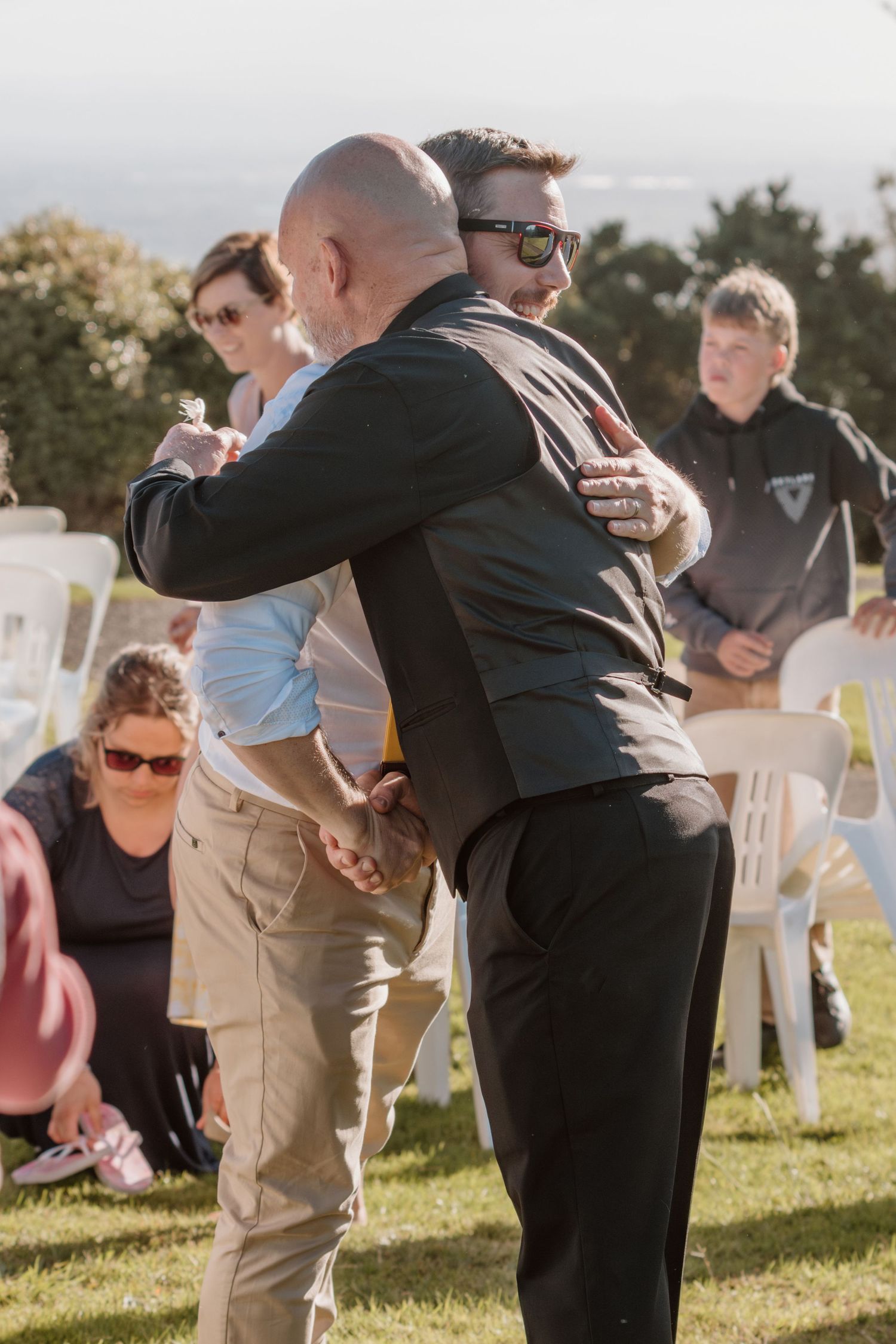 Two groomsmen share an emotional hug during an outdoor wedding ceremony on a sunny day with white chairs in background.