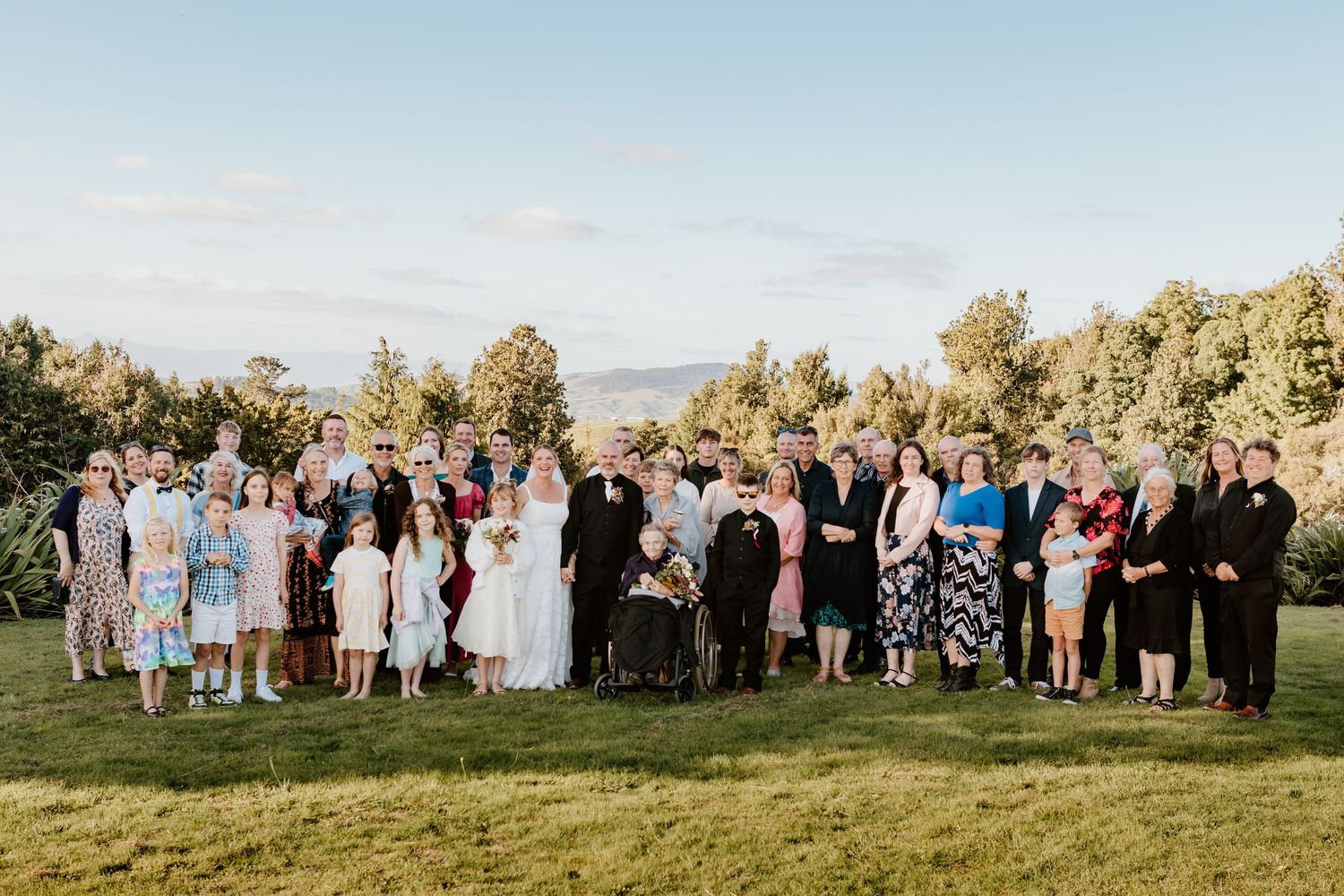 Wedding party and guests stand together on a grassy hilltop during golden hour with mountains in the background.