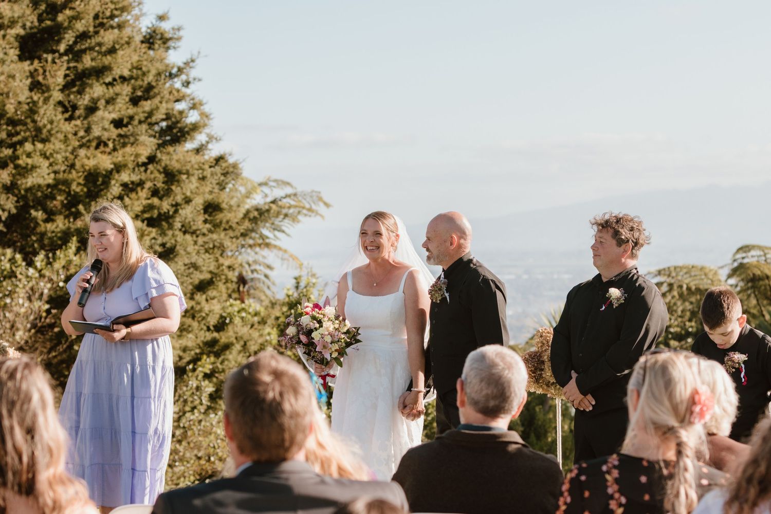 Scenic outdoor wedding ceremony at sunset with guests standing under evergreen trees.