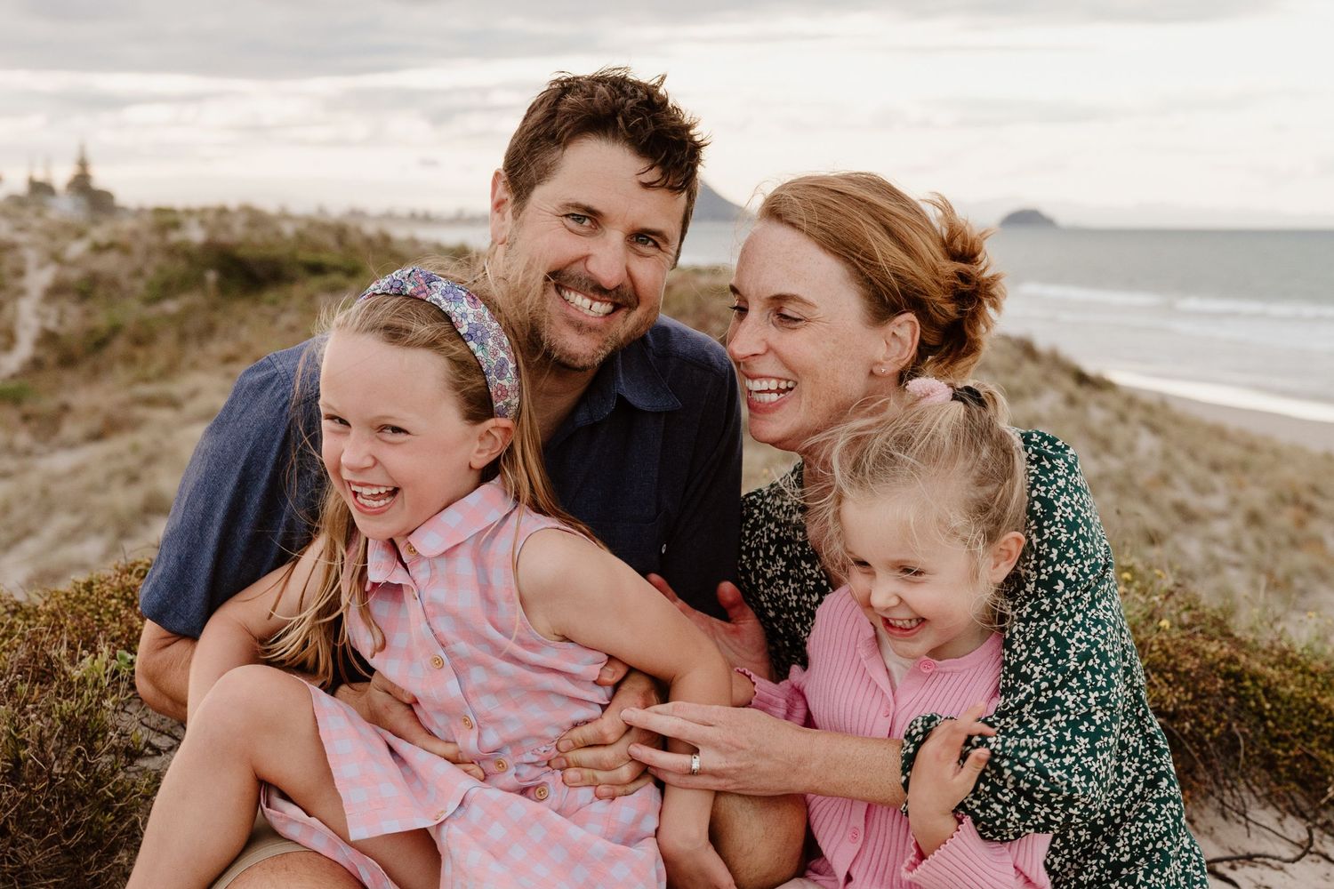 A joyful family shares loving laughter together while sitting on a coastal hill overlooking the beach at sunset.