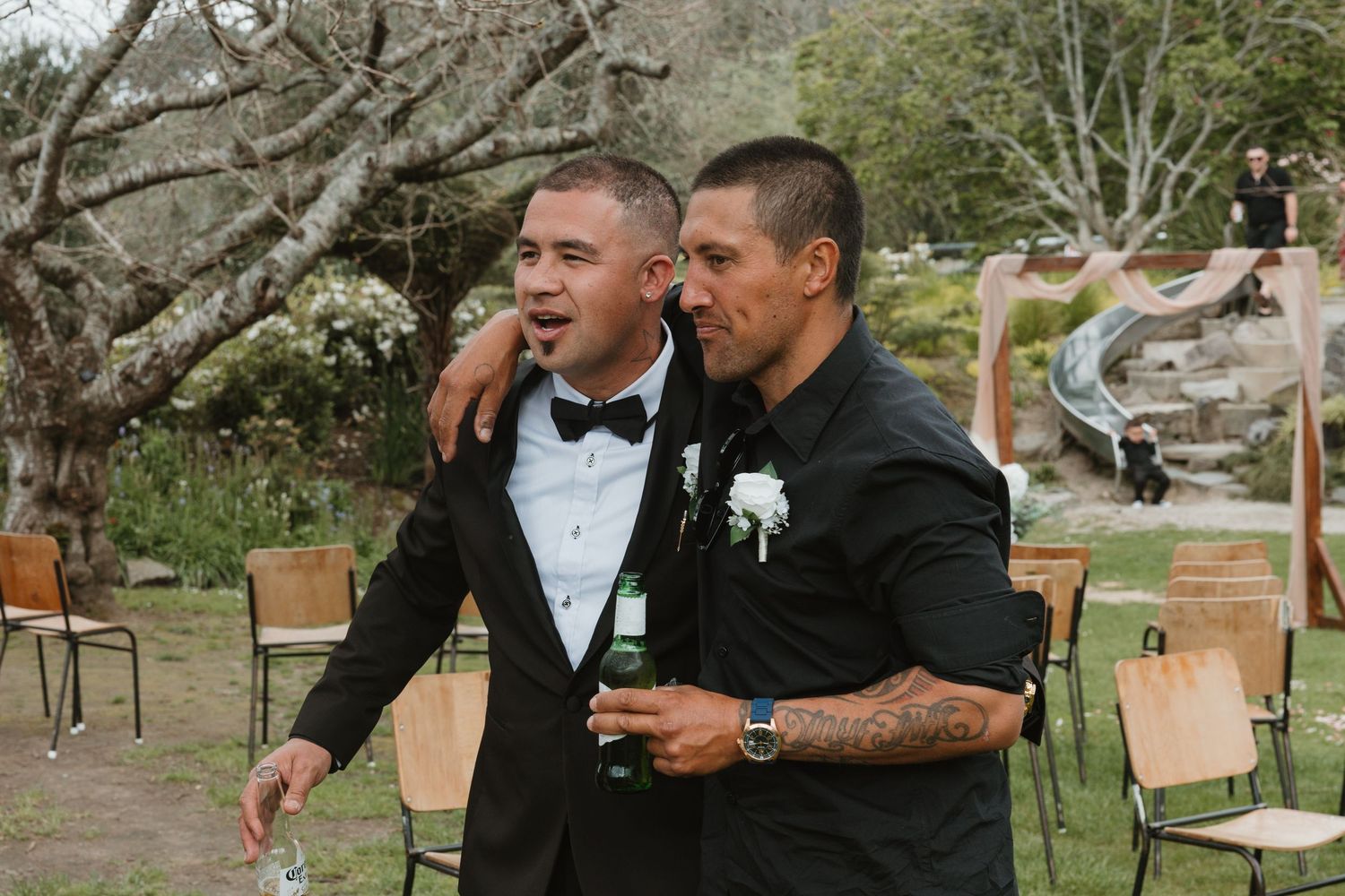 Two men in black tuxedos share a candid moment at an outdoor wedding ceremony setup with wooden chairs.