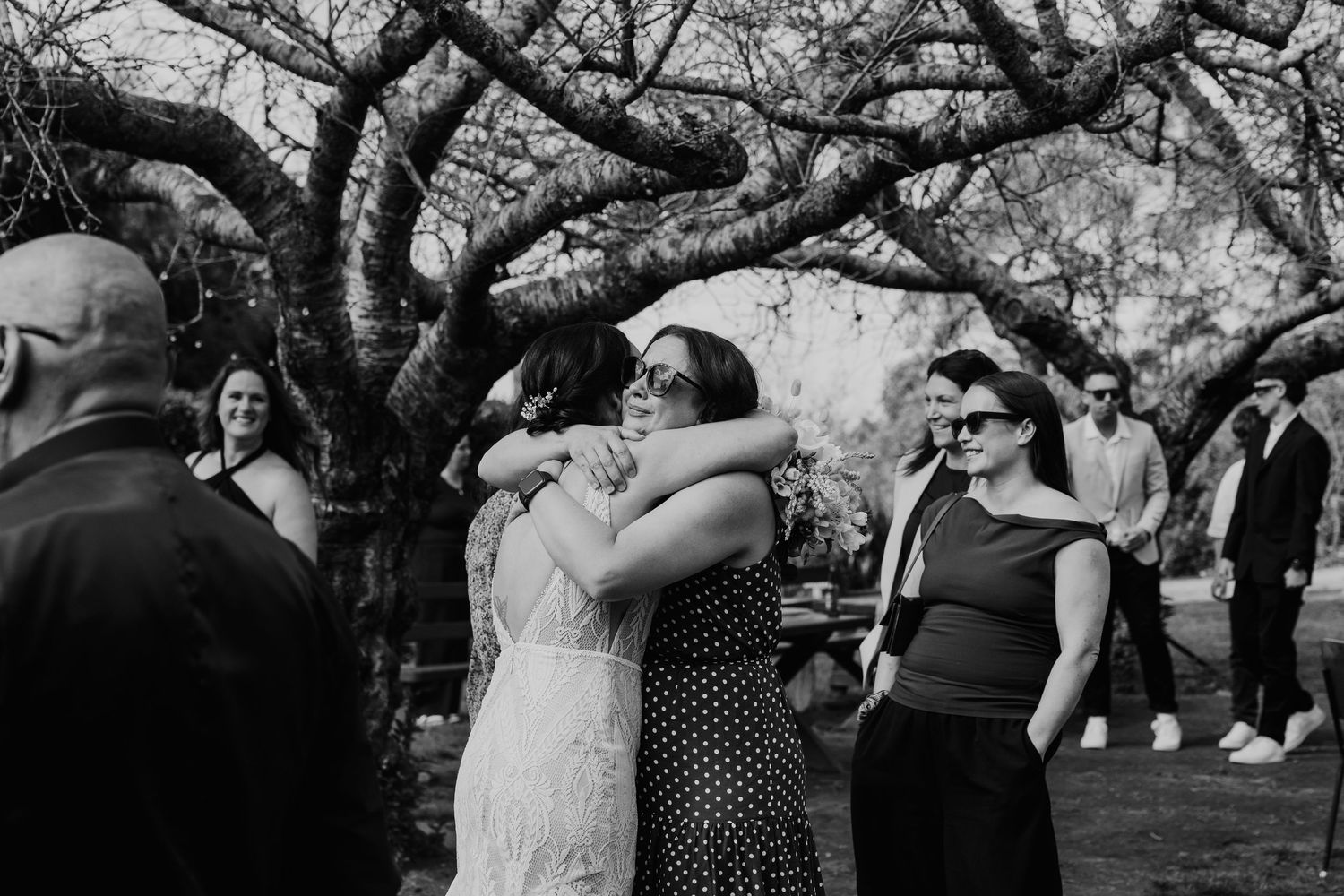 Wedding guests gather outdoors under sprawling trees during a daytime celebration ceremony.
