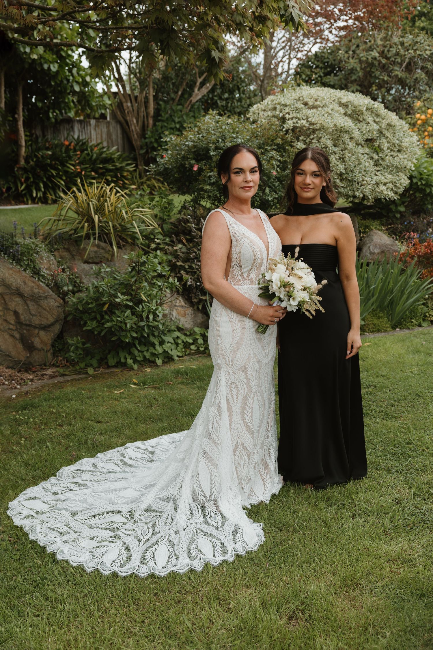 Two people pose together on a lawn, one wearing a white lace wedding gown with a long train and one in a black dress.