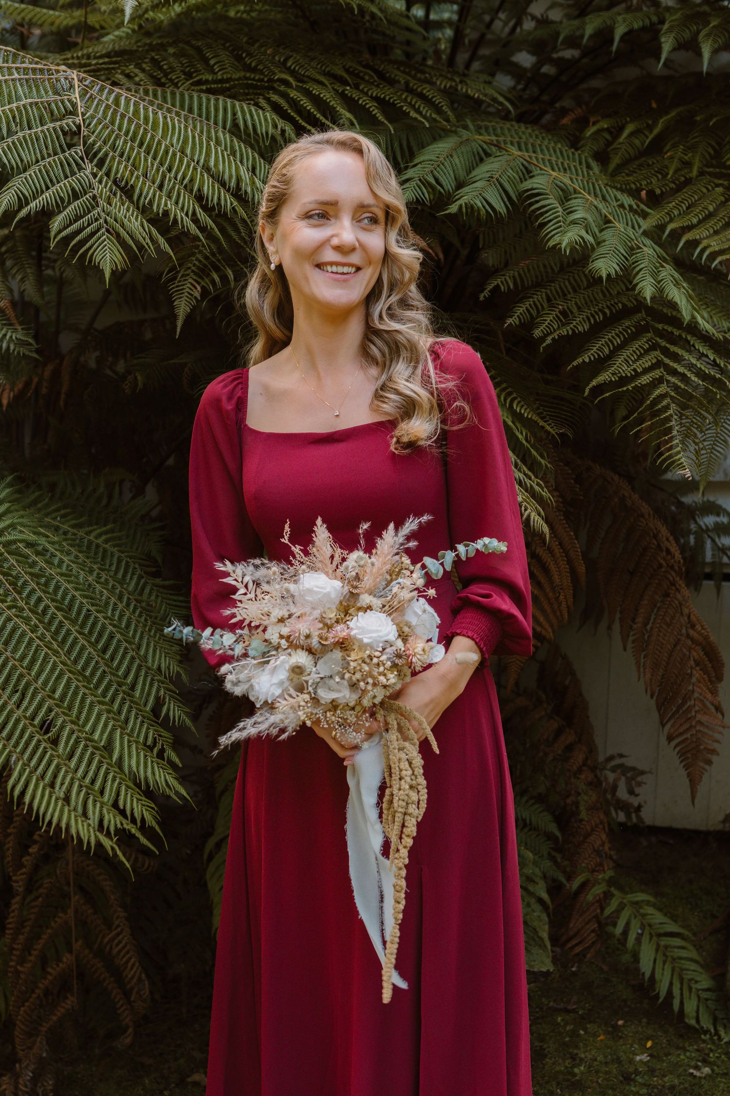 A person in a burgundy dress holds a rustic dried floral bouquet against a backdrop of lush green ferns.