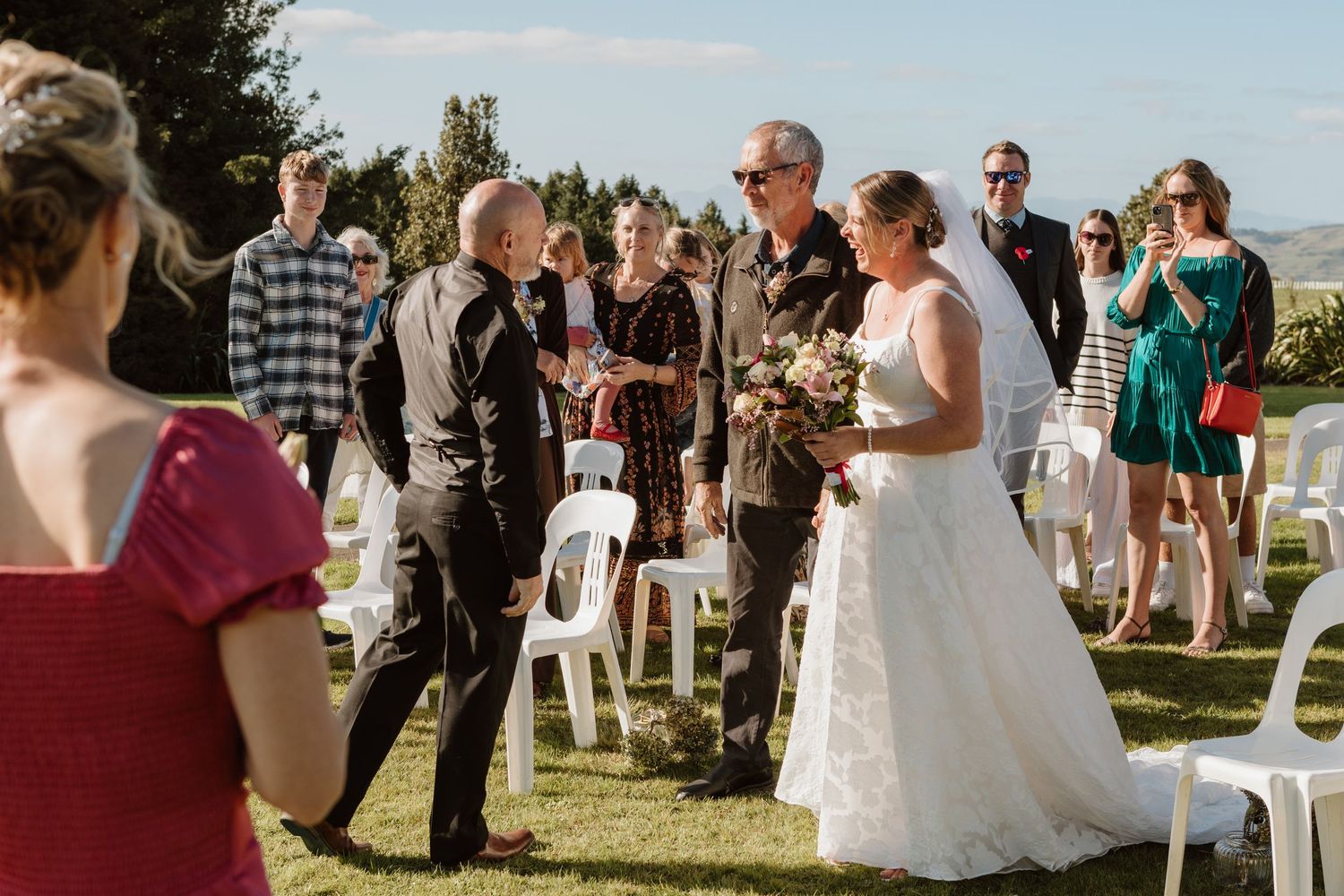 A beautiful outdoor wedding ceremony takes place on a grassy lawn with a scenic mountain backdrop on a sunny day.